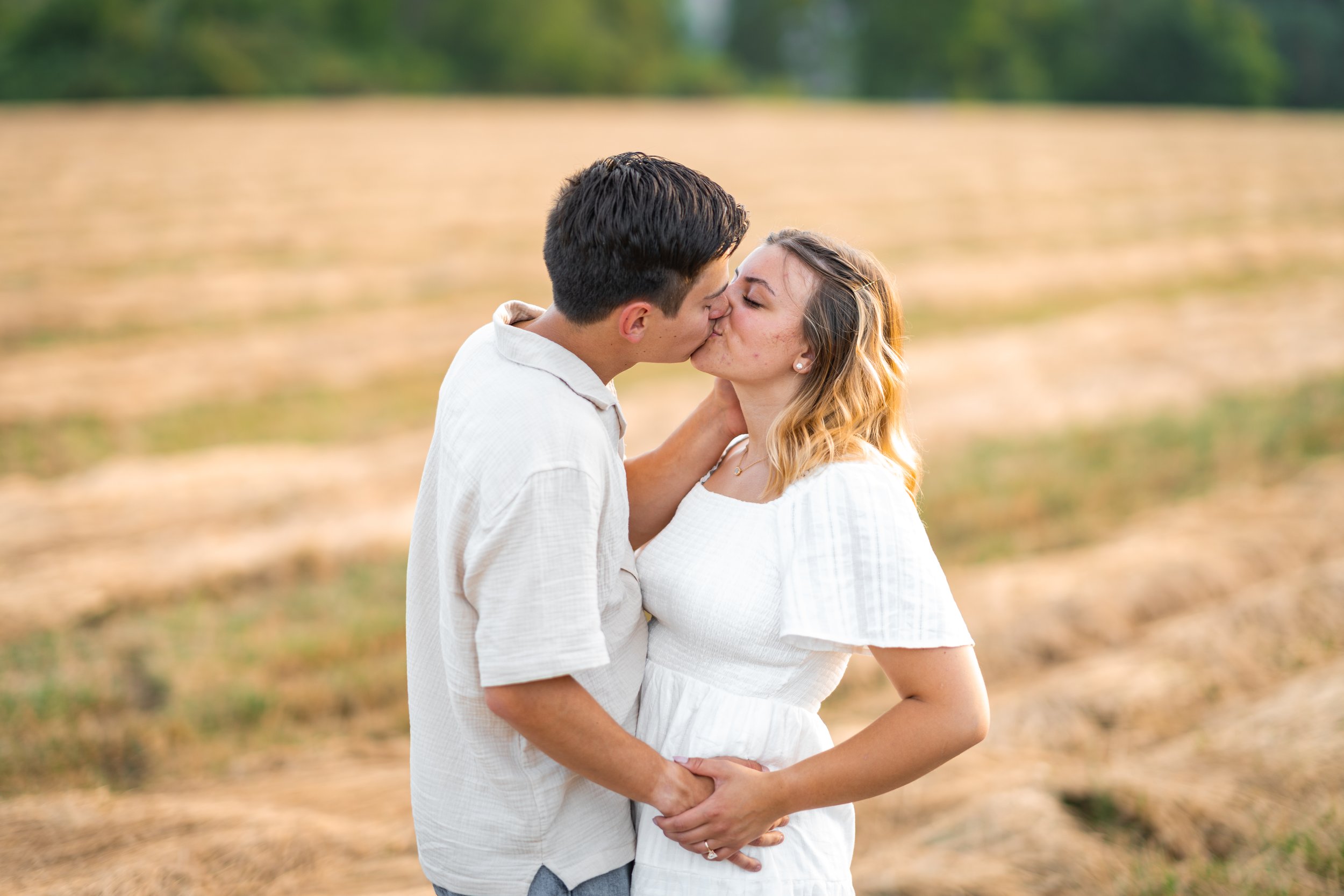 A young couple in love sharing a kiss outdoors in a field, with green trees in the background.