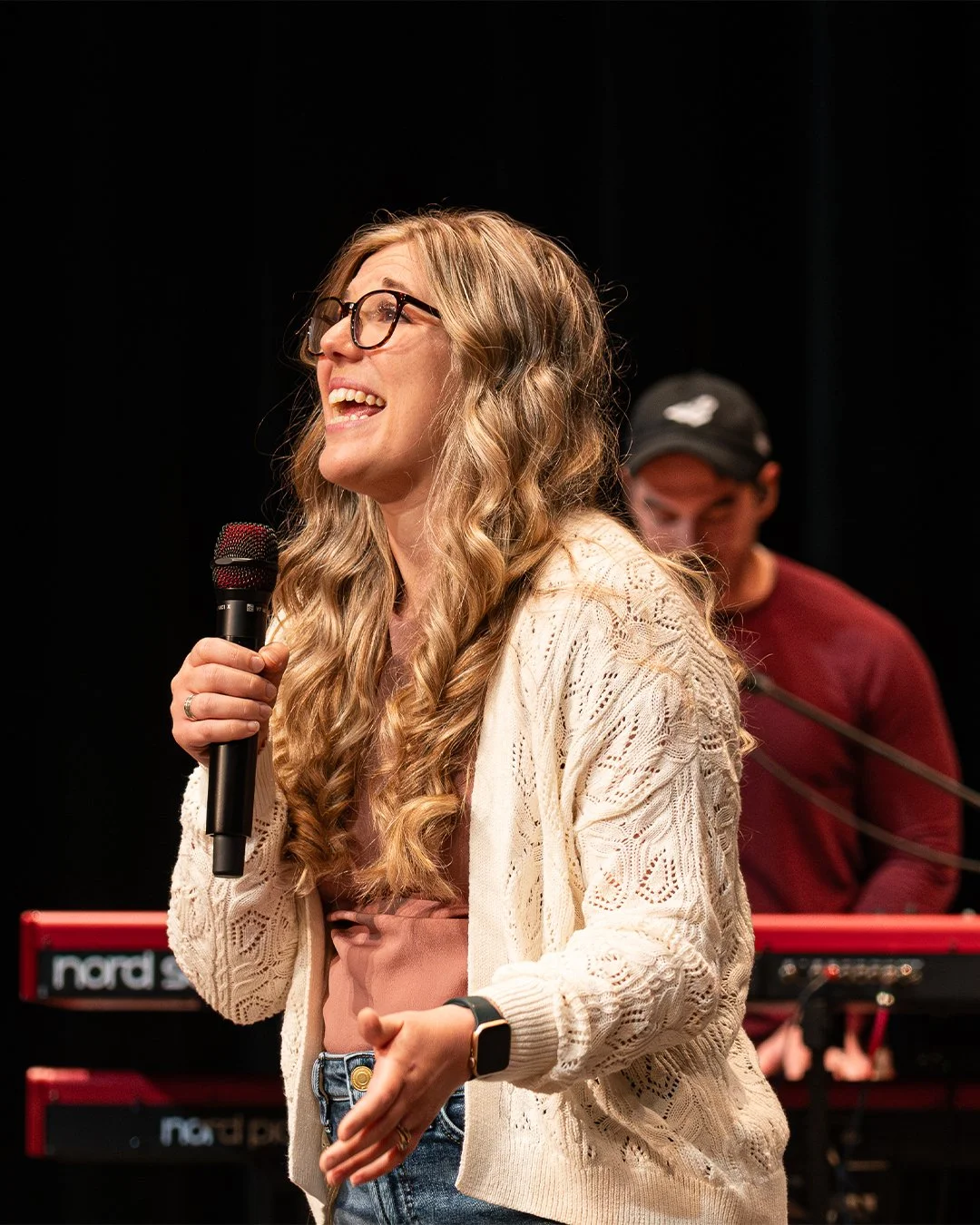 A woman with long wavy blonde hair, glasses, and a beige knit cardigan is smiling and holding a microphone, standing on stage with a man playing a keyboard in the background.
