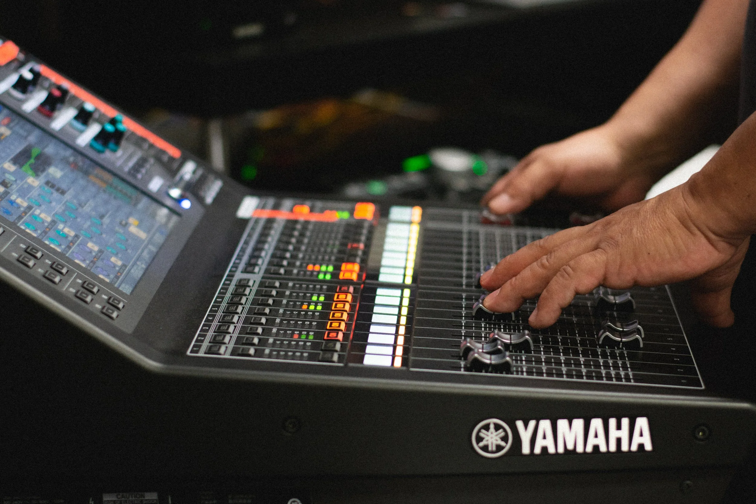 Close-up of a Yamaha mixing console with multiple colorful sliders, knobs, and a touchscreen display, and a person's hand adjusting controls.