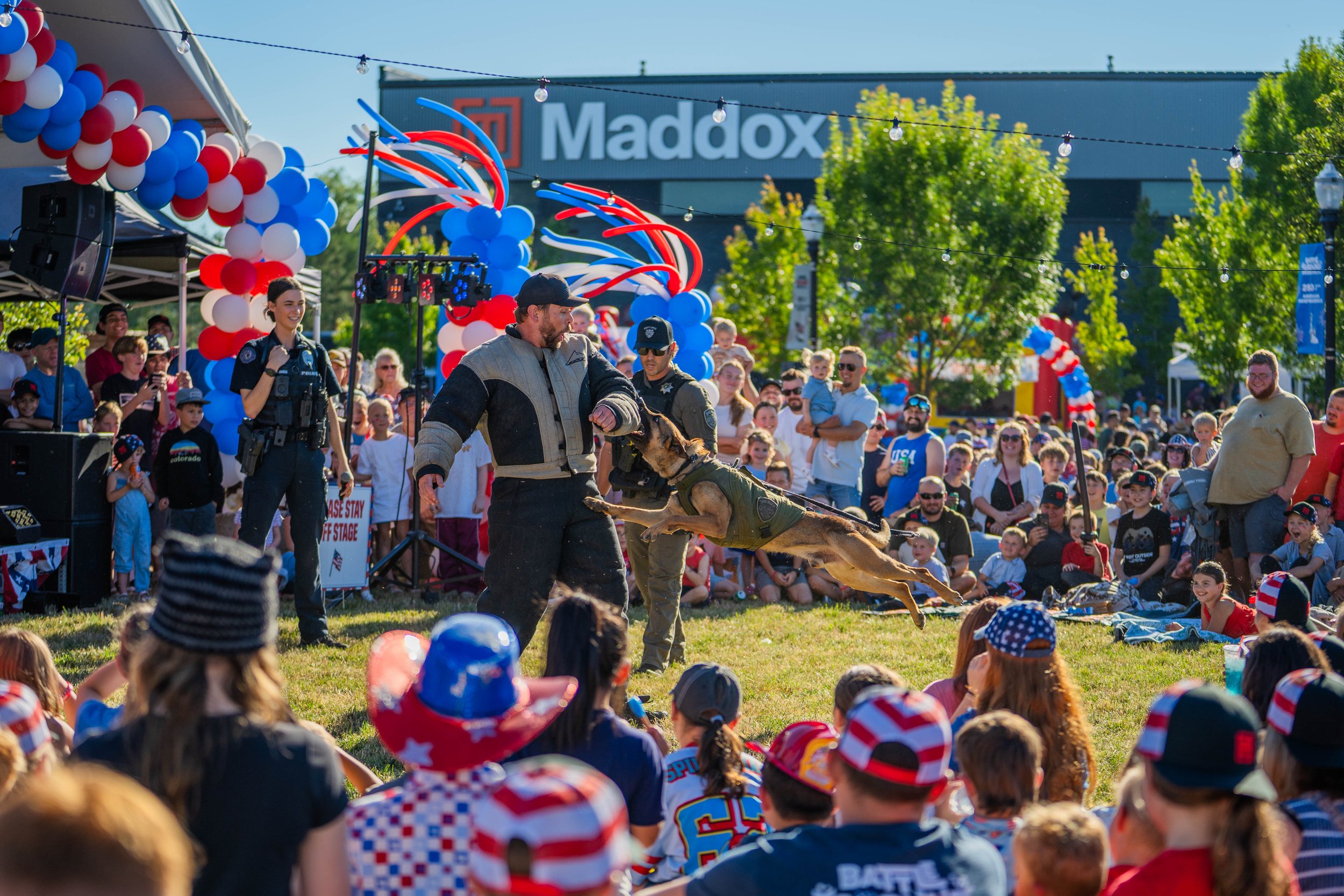 Crowd gathered at an outdoor event with patriotic decorations, including balloons in red, white, and blue, and a large billboard with the logo 'Maddox' in the background. Police officers and a man with a German Shepherd dog are participating in a dem