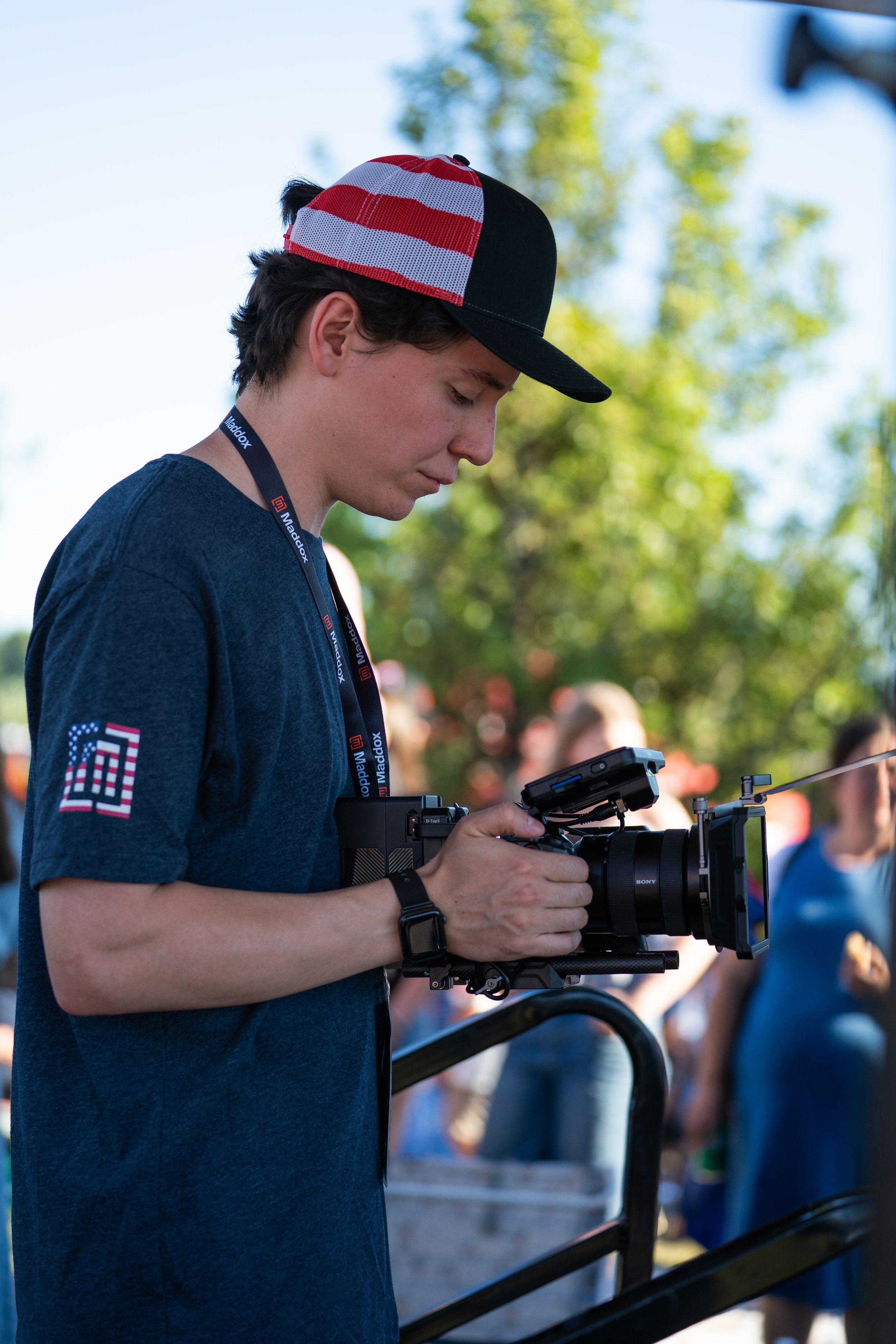 Young man with dark hair wearing a red, white, and black cap, a blue shirt with an American flag patch on the sleeve, and a camera strap around his neck, operating a professional camera rig outdoors in daytime, with a background of trees and people.
