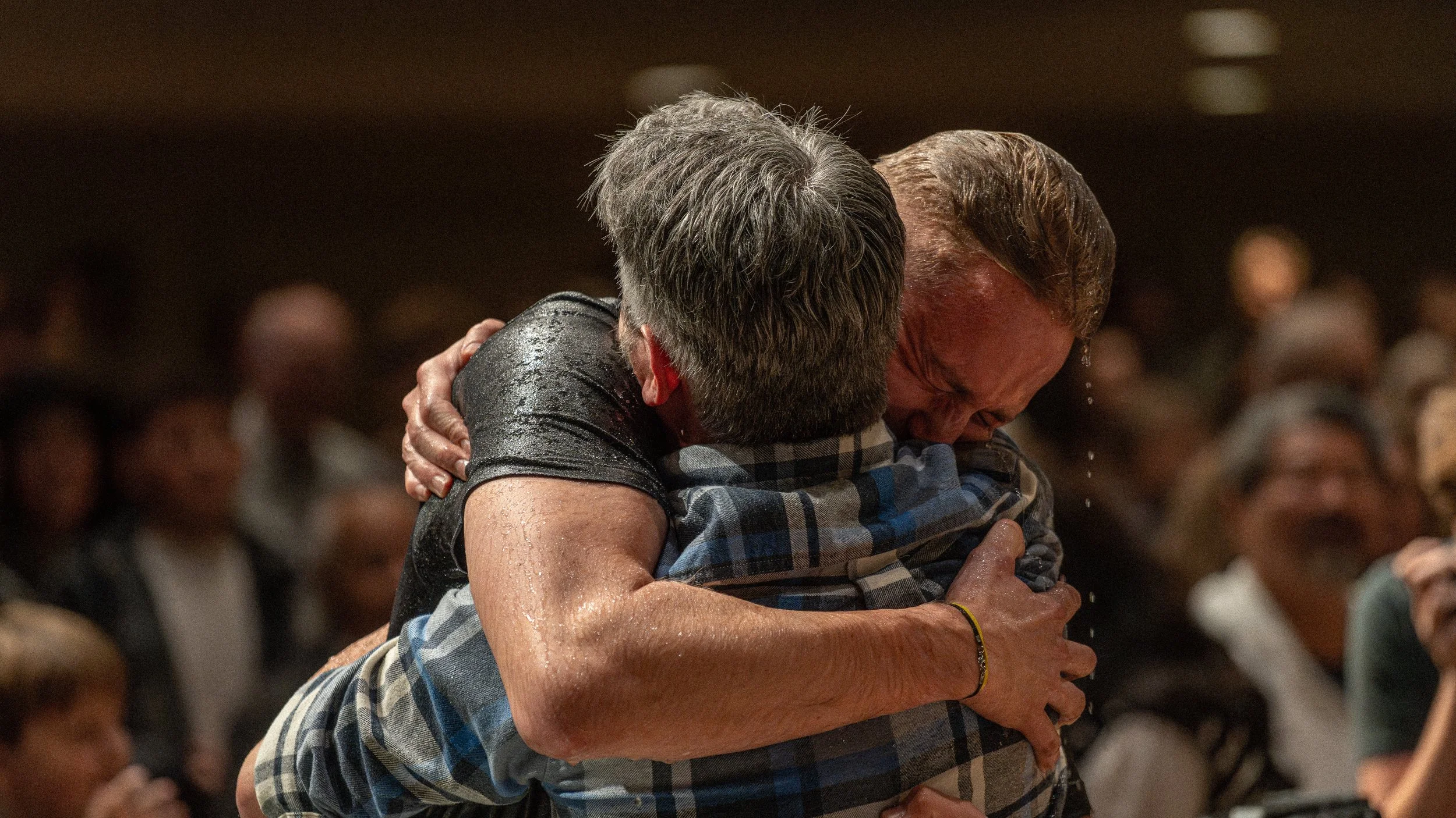 Two men embracing in a hug, visibly emotional, in a crowded indoor setting.