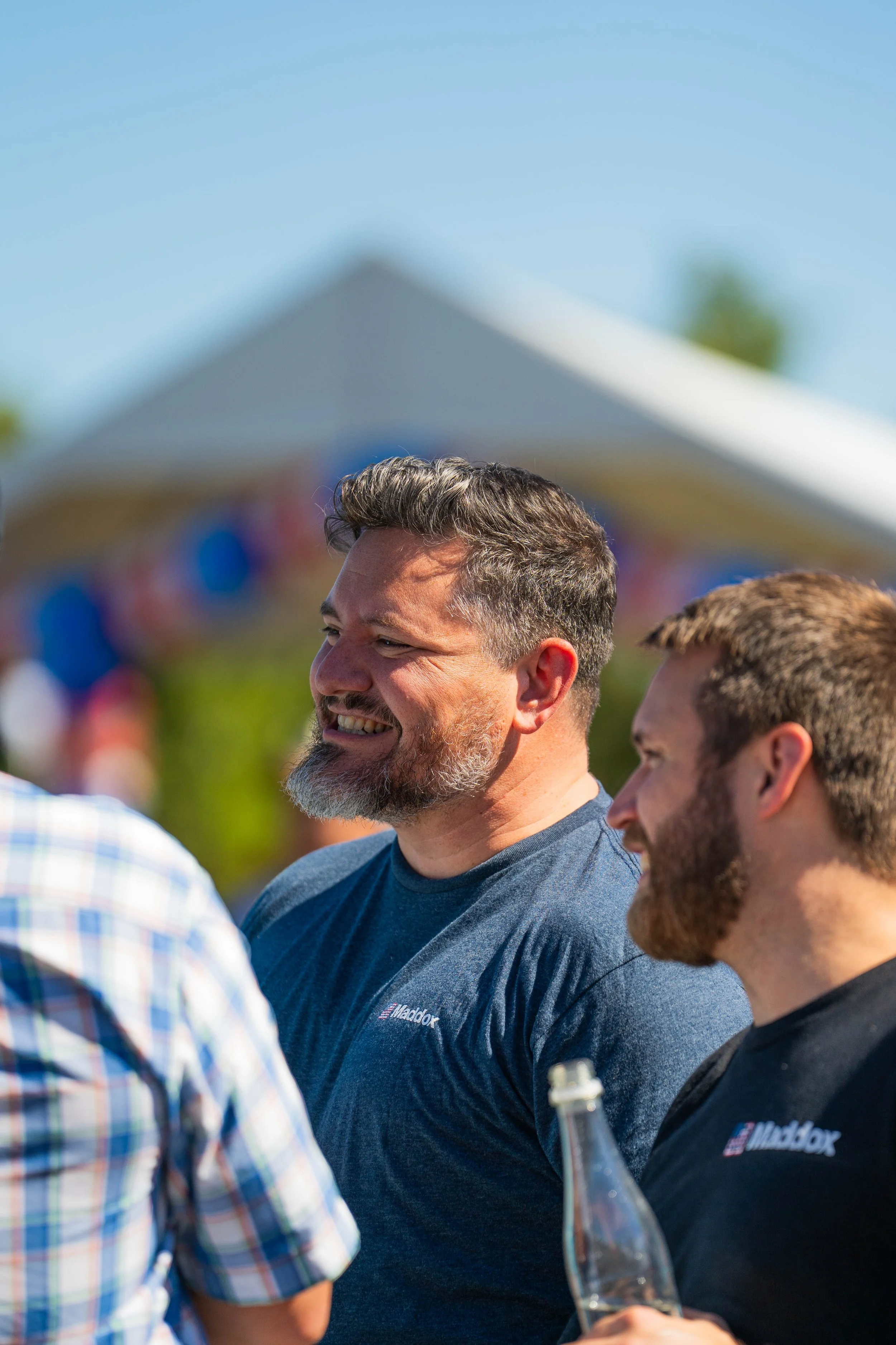 Group of men outdoors during daytime, talking and smiling, with a tent and trees in the background.
