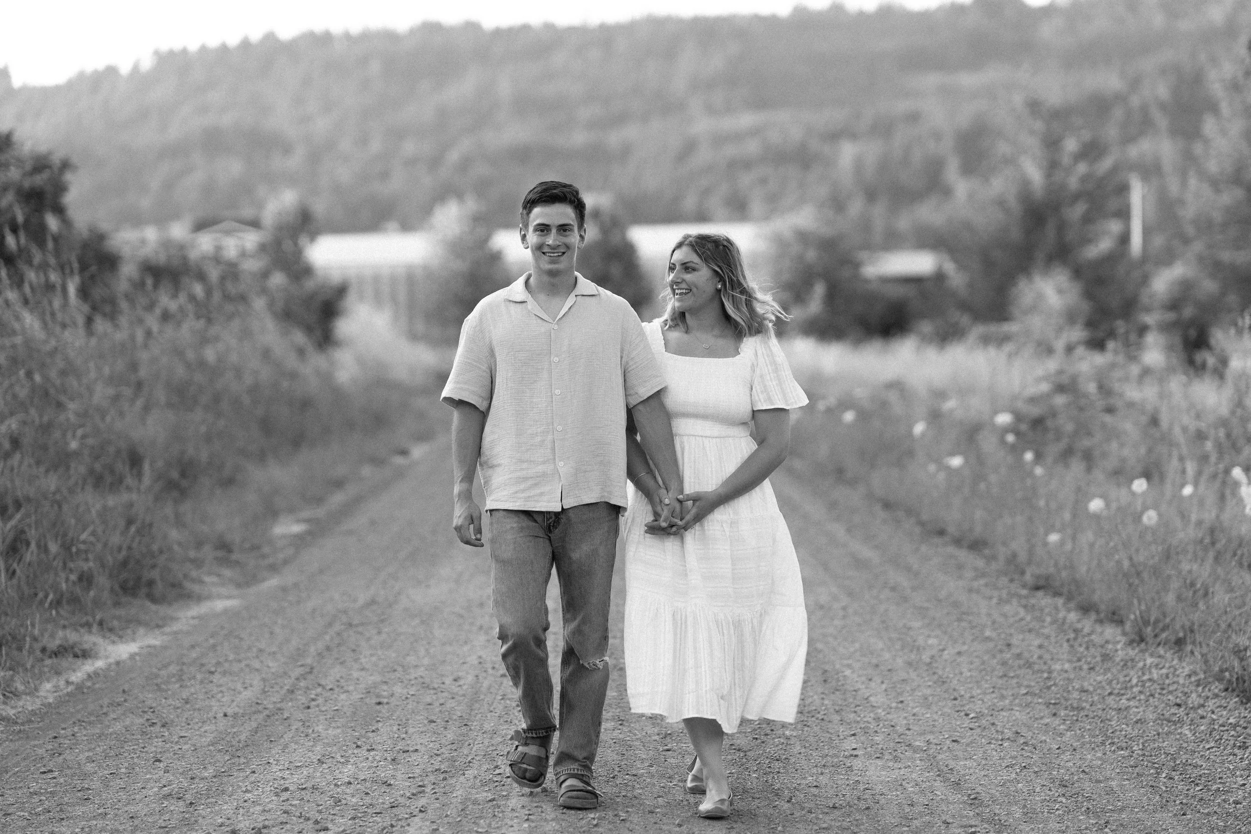 A young couple walking hand-in-hand down a rural dirt road surrounded by greenery and hills, smiling and enjoying each other's company.