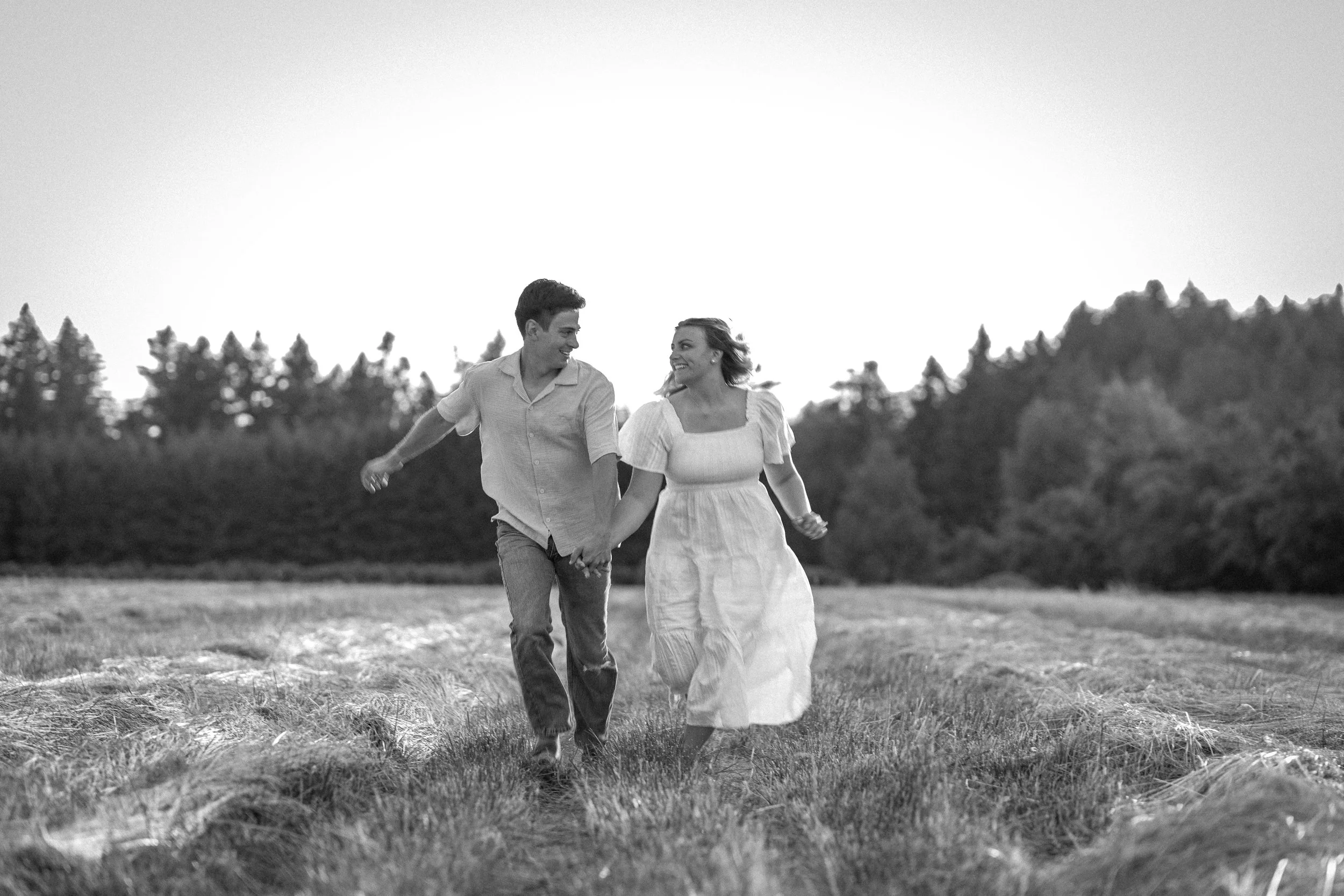 A black and white photo of a young couple holding hands while walking through a grassy field, smiling at each other.