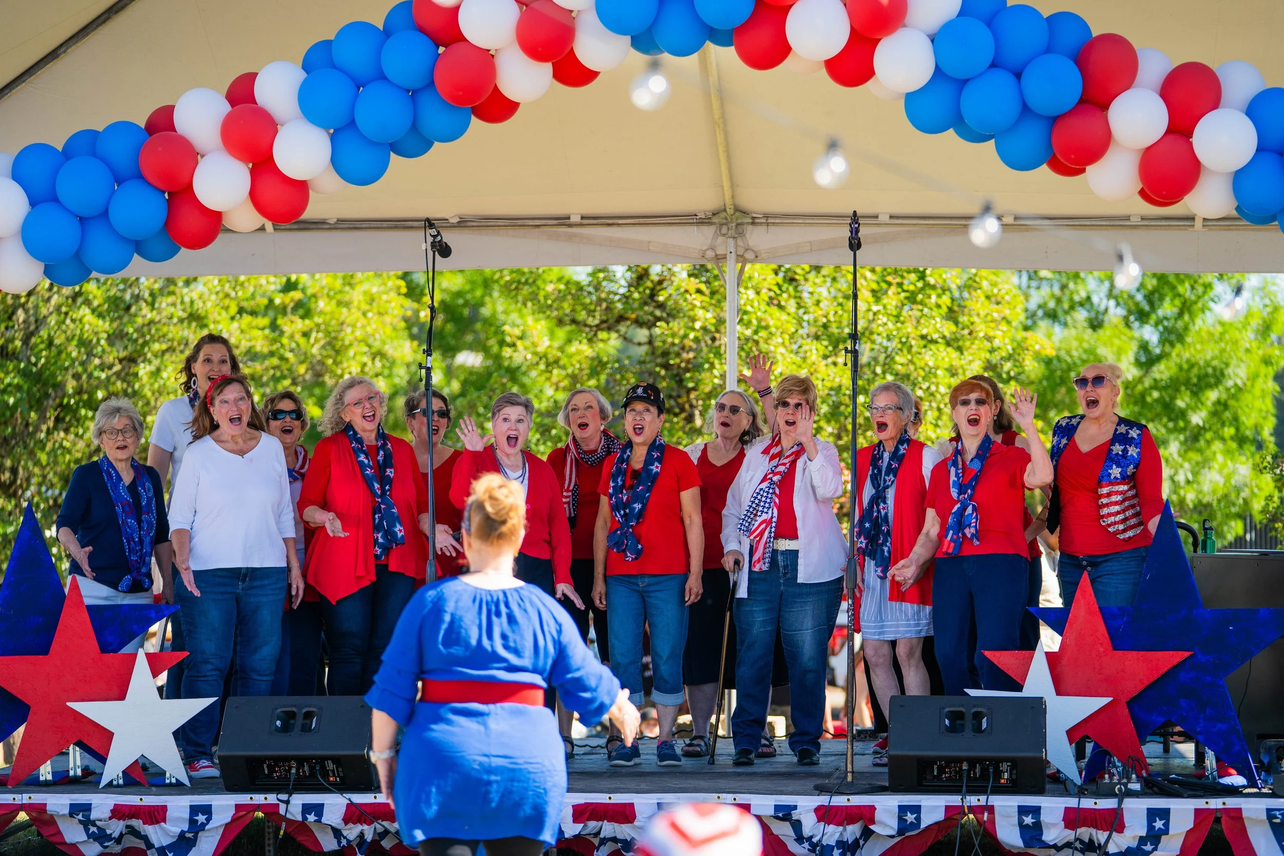 A group of women singing on an outdoor stage decorated with red, white, and blue stars and bunting, with balloons in red, white, and blue forming an arch overhead, during a patriotic celebration.