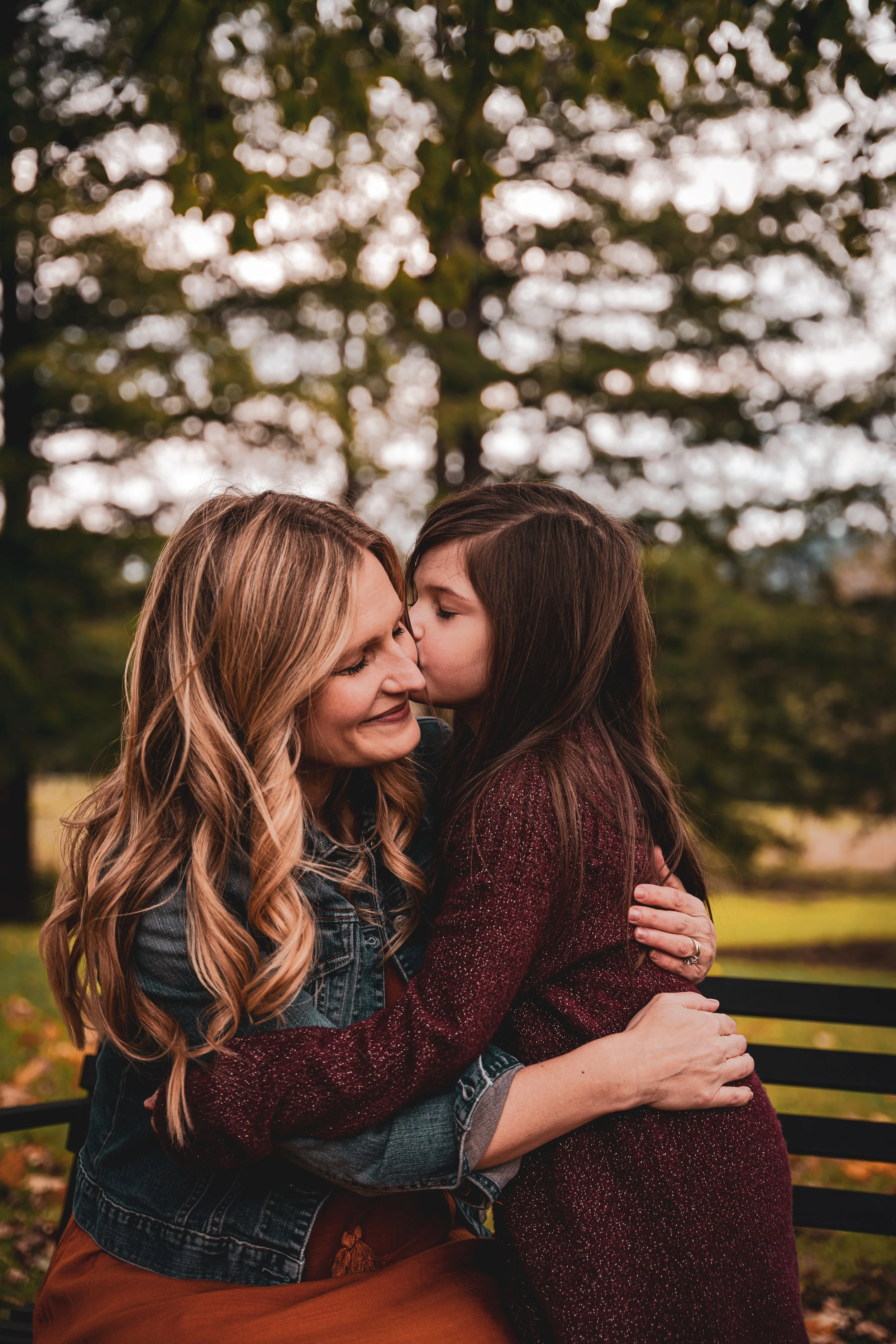 A woman with long, wavy blonde hair, wearing a denim jacket, hugs a woman with straight, dark brown hair, wearing a maroon sweater, as they share a tender moment outside on a park bench with trees in the background.