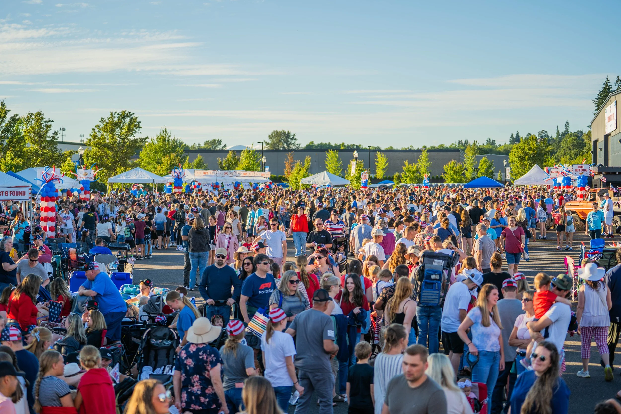 A large crowd of people gathered outdoors during a patriotic event, with many wearing red, white, and blue clothing and accessories, under a clear sky.