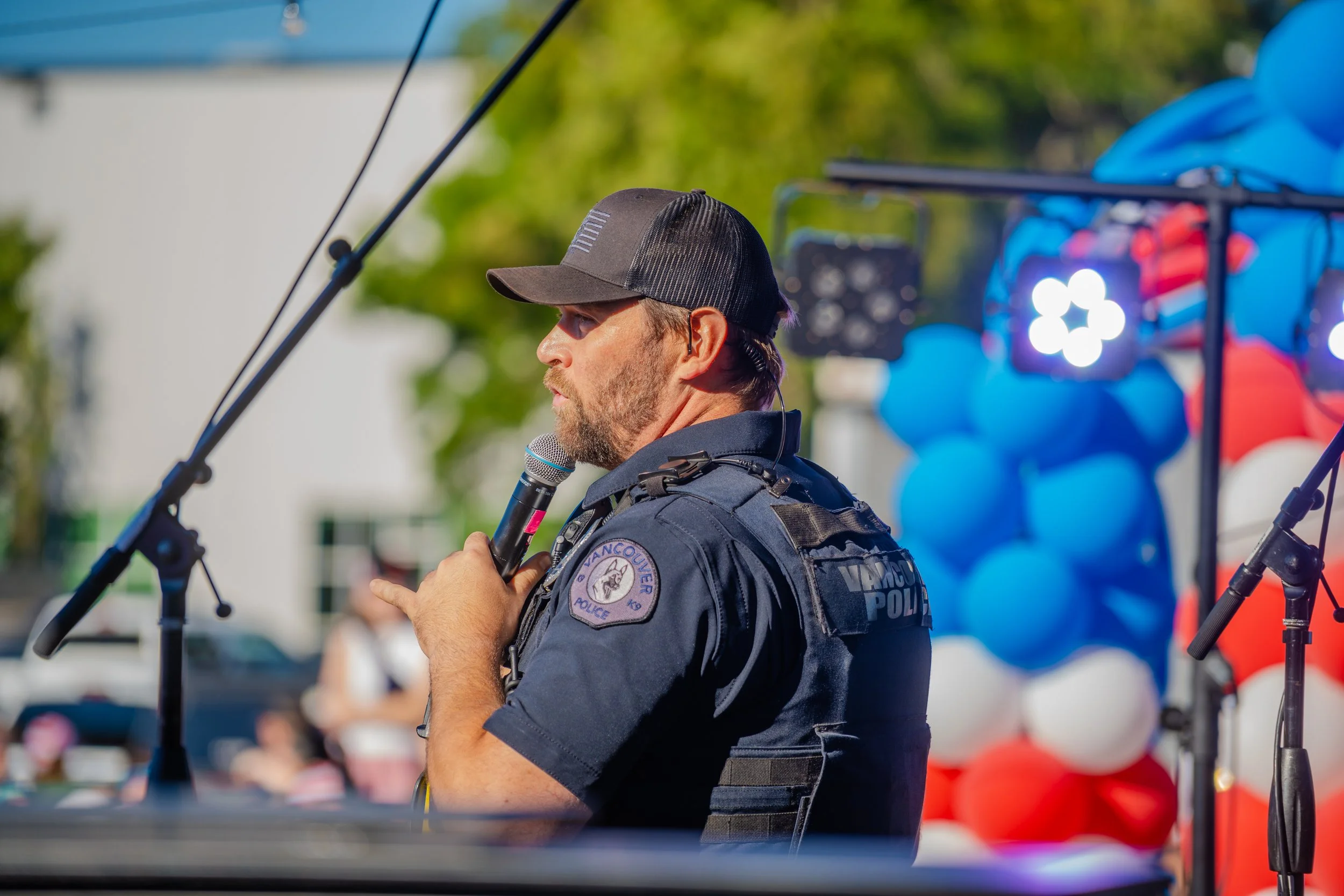 A police officer speaking into a microphone during an outdoor event, with a colorful inflatable structure in the background.