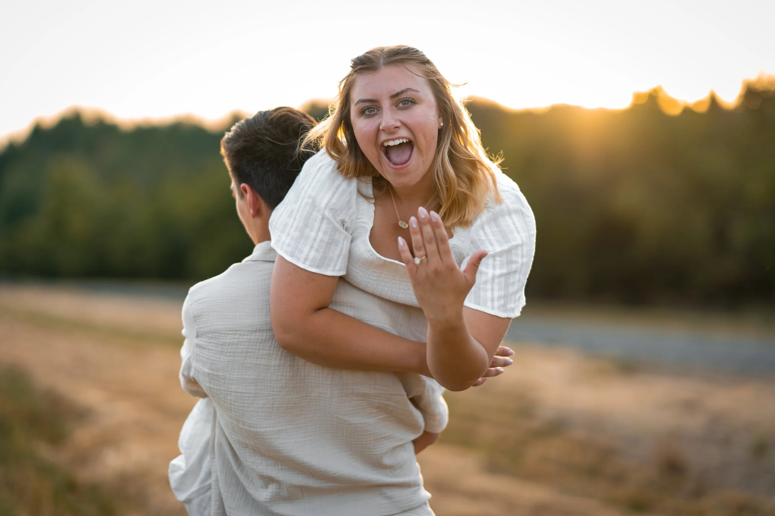 A woman with blonde hair and a white shirt is smiling and waving while being carried on the back of a man, with a blurred outdoor background and warm lighting.