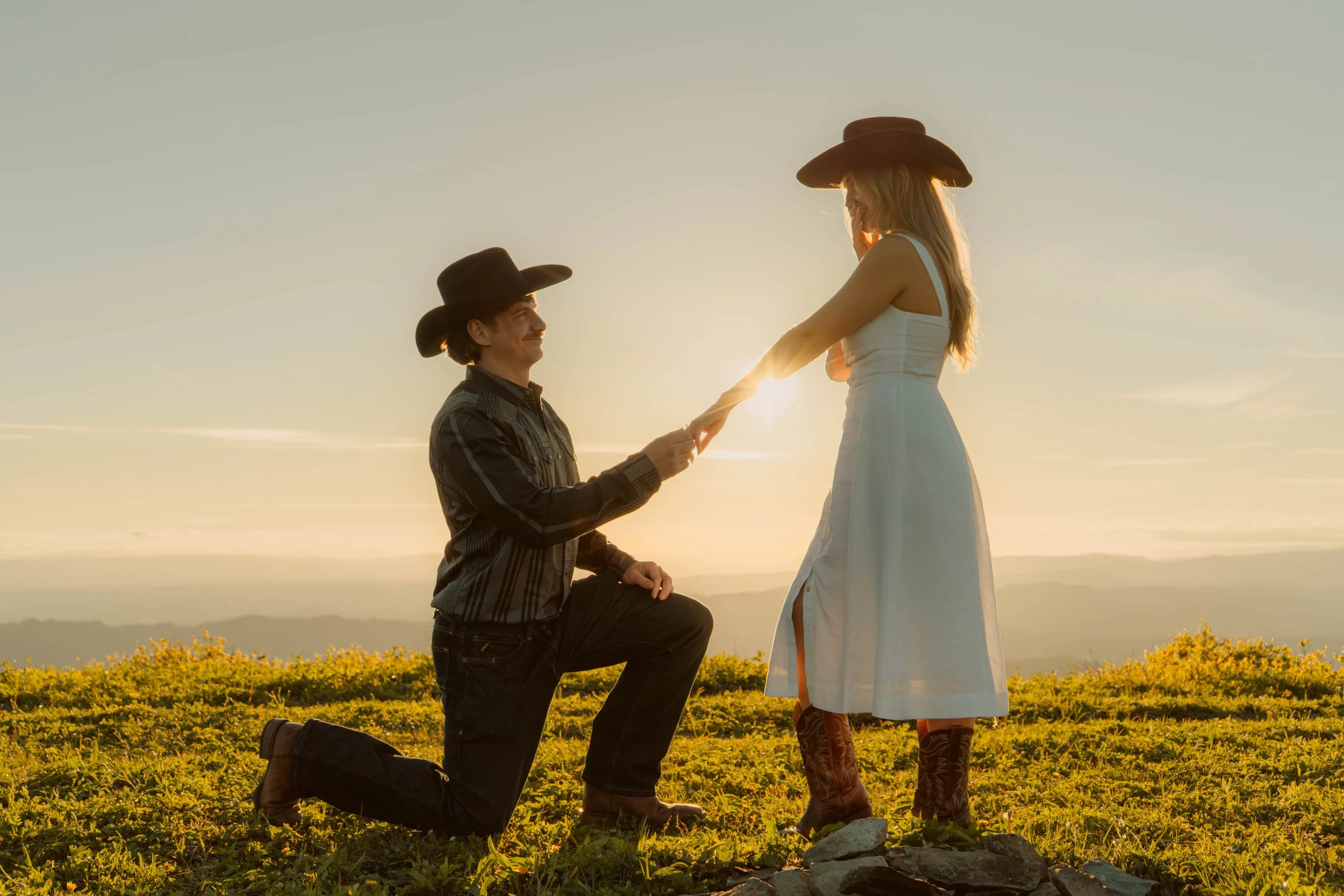 A man on one knee holding a woman's hand during a proposal, set against a sunset in an open field with yellow flowers and distant mountains.
