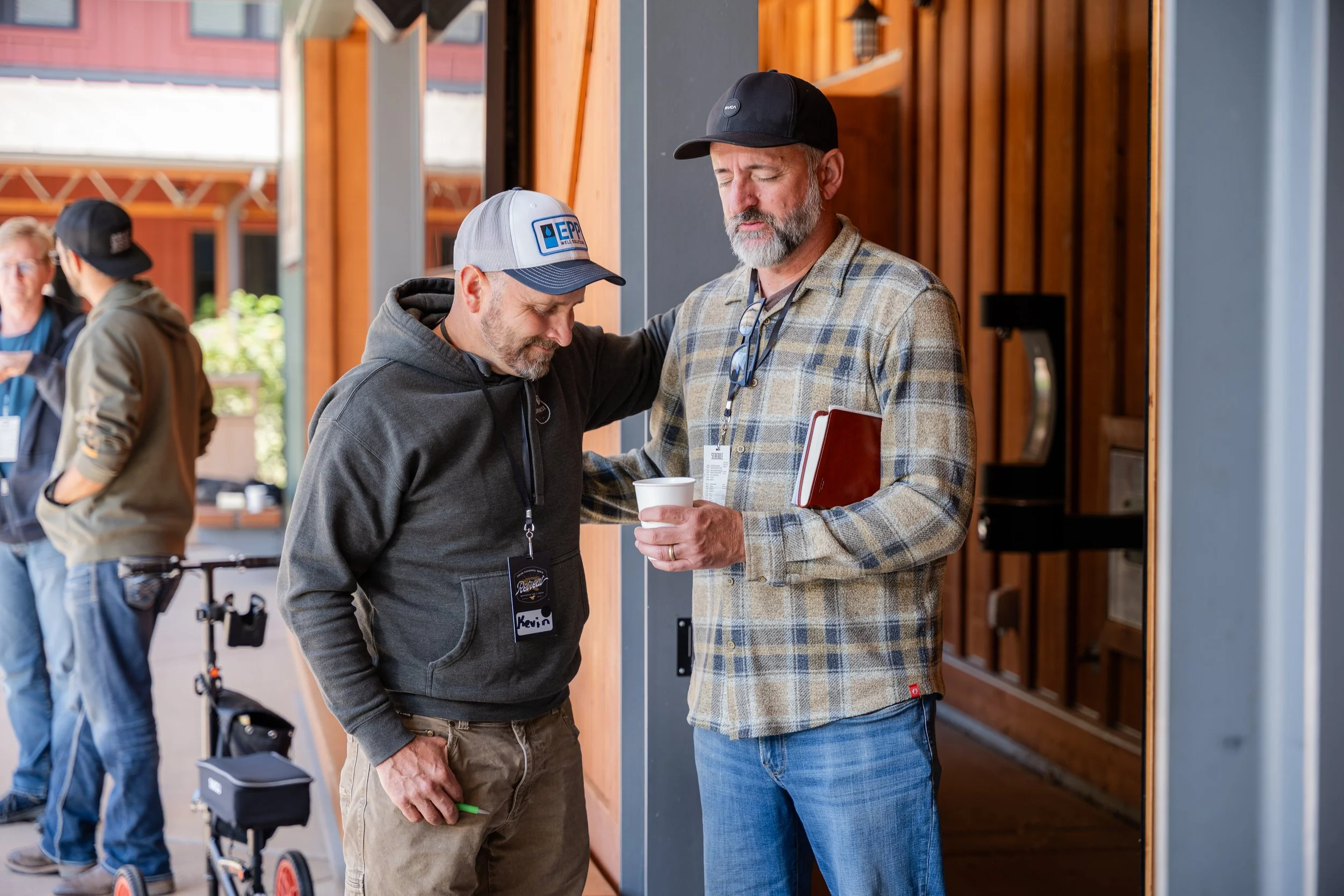 Two men are talking closely at an event or conference, one of them is holding a paper cup. One man wears a gray hoodie with a name tag that reads 'Kevin' and a baseball cap, the other wears a beige plaid jacket, a black baseball cap, and sunglasses h