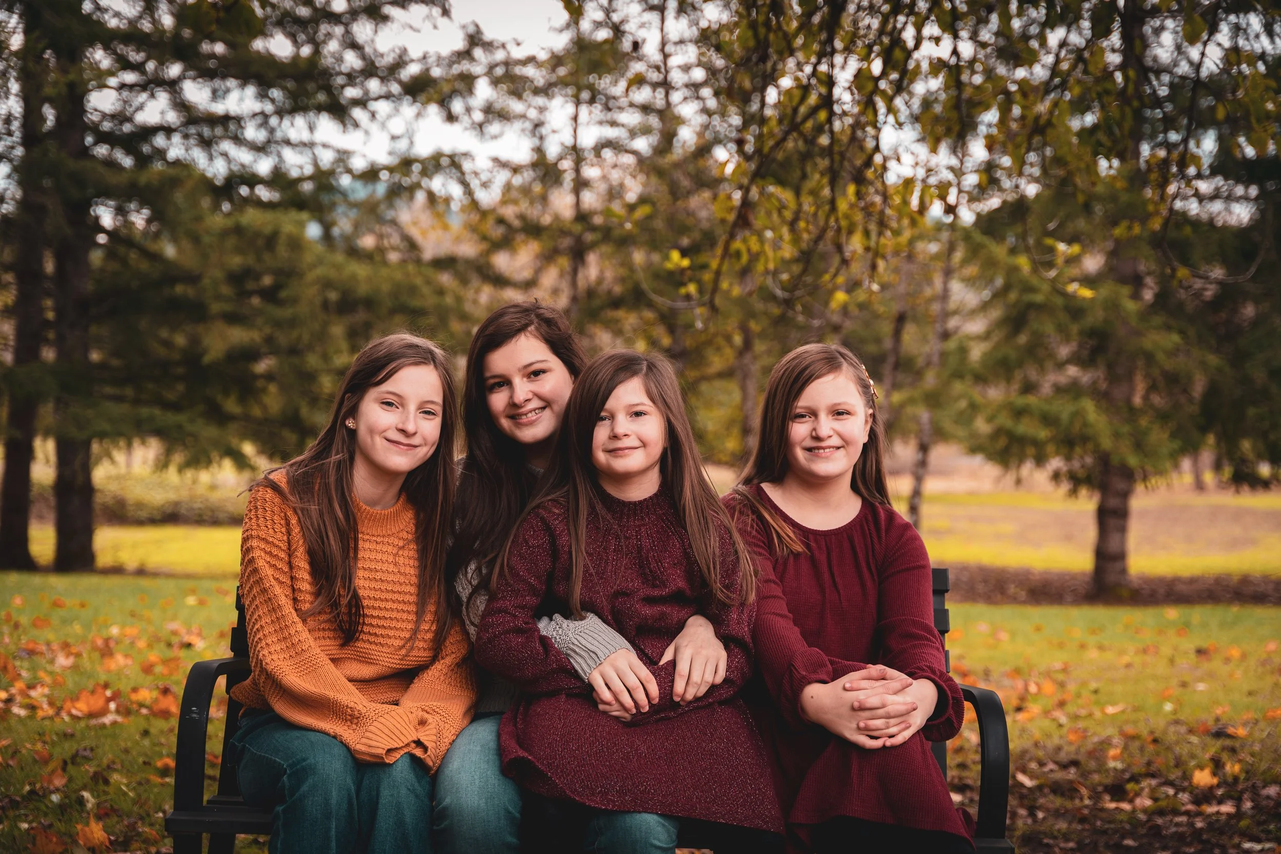 Four smiling girls sitting on a park bench outdoors during fall, surrounded by trees with autumn leaves. They are dressed in warm sweaters and have light to medium brown hair.