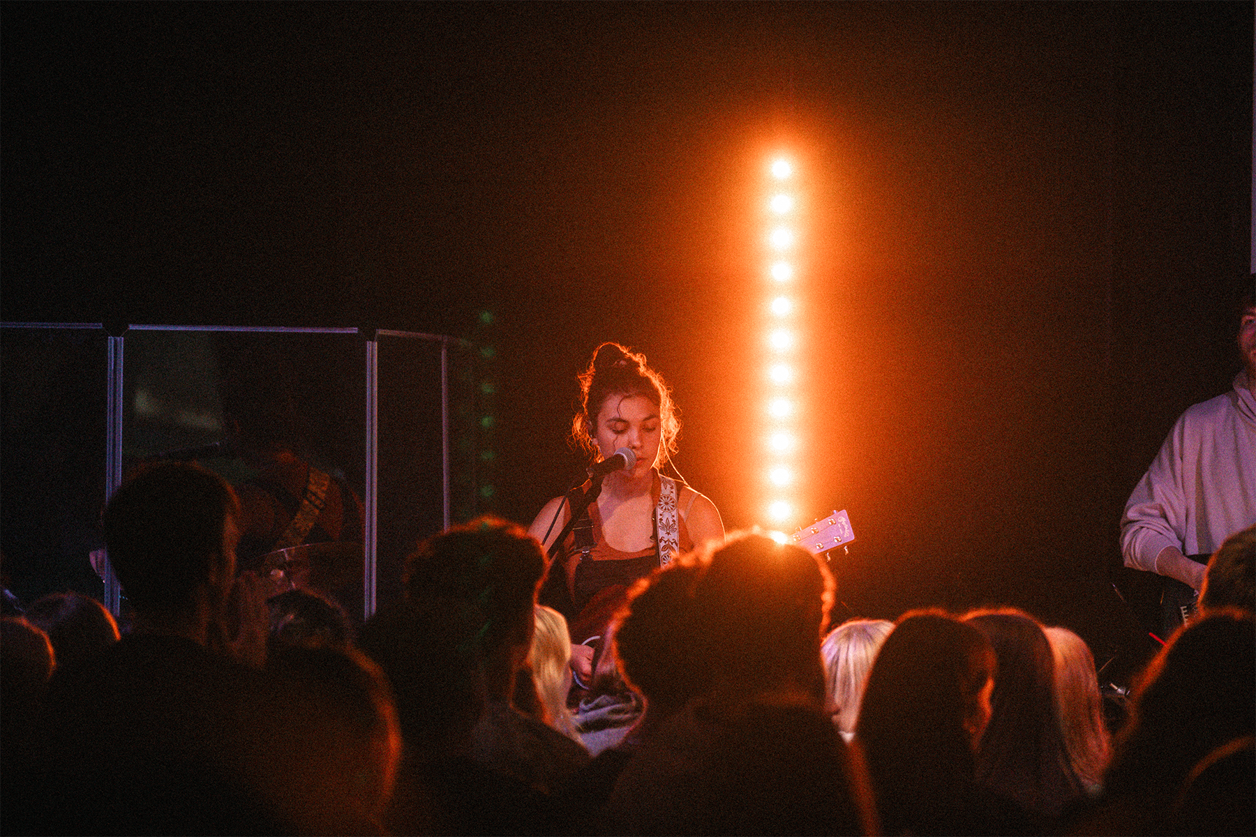 A young woman with her hair in a messy bun sings into a microphone while playing an electric guitar at a live concert. Audience members are visible in the foreground with stage lighting behind her creating an orange glow.