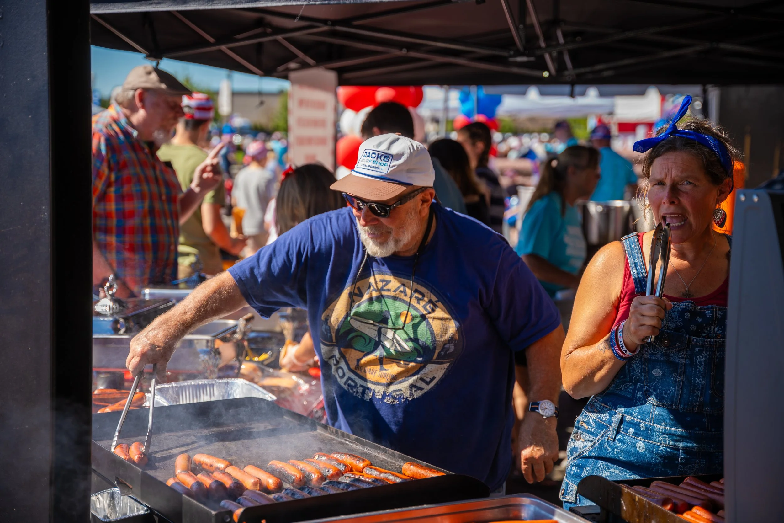People gather at an outdoor food stand grilling hot dogs and sausages under a canopy, with balloons and a crowd of vendors and attendees visible in the background.