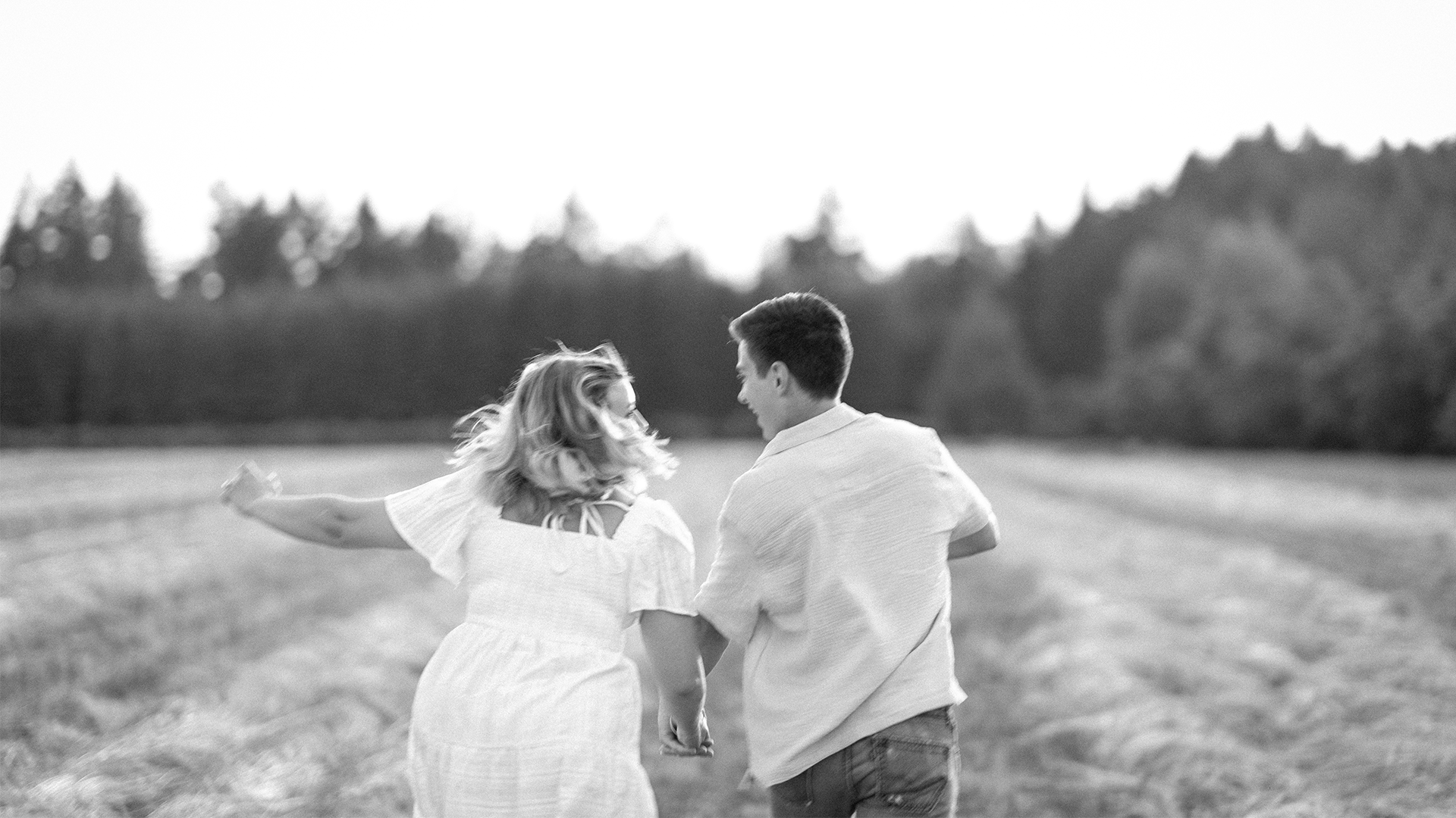 A black-and-white photo of a woman and a man walking in a field, holding hands and smiling at each other, with trees in the background.