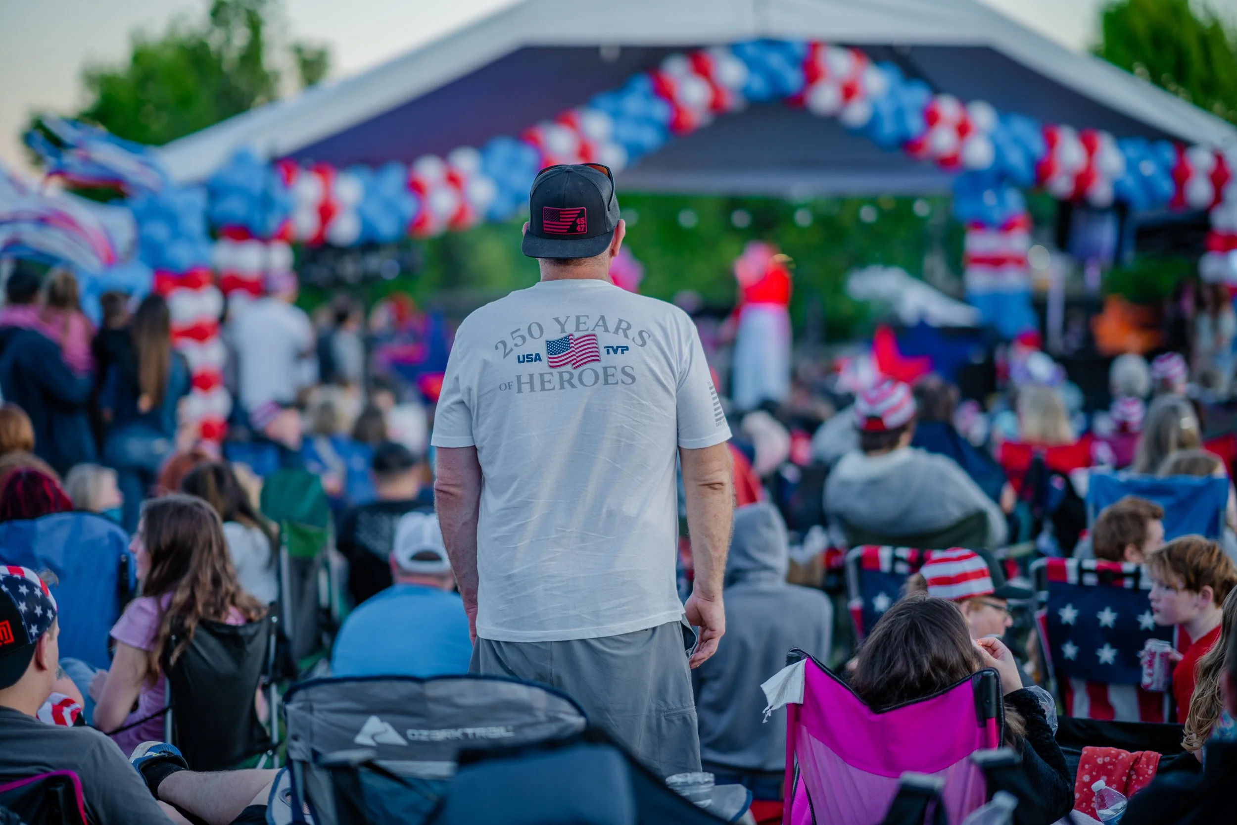 A man wearing a cap and a T-shirt with patriotic text and flag, standing in front of a crowd at an outdoor event with patriotic decorations.