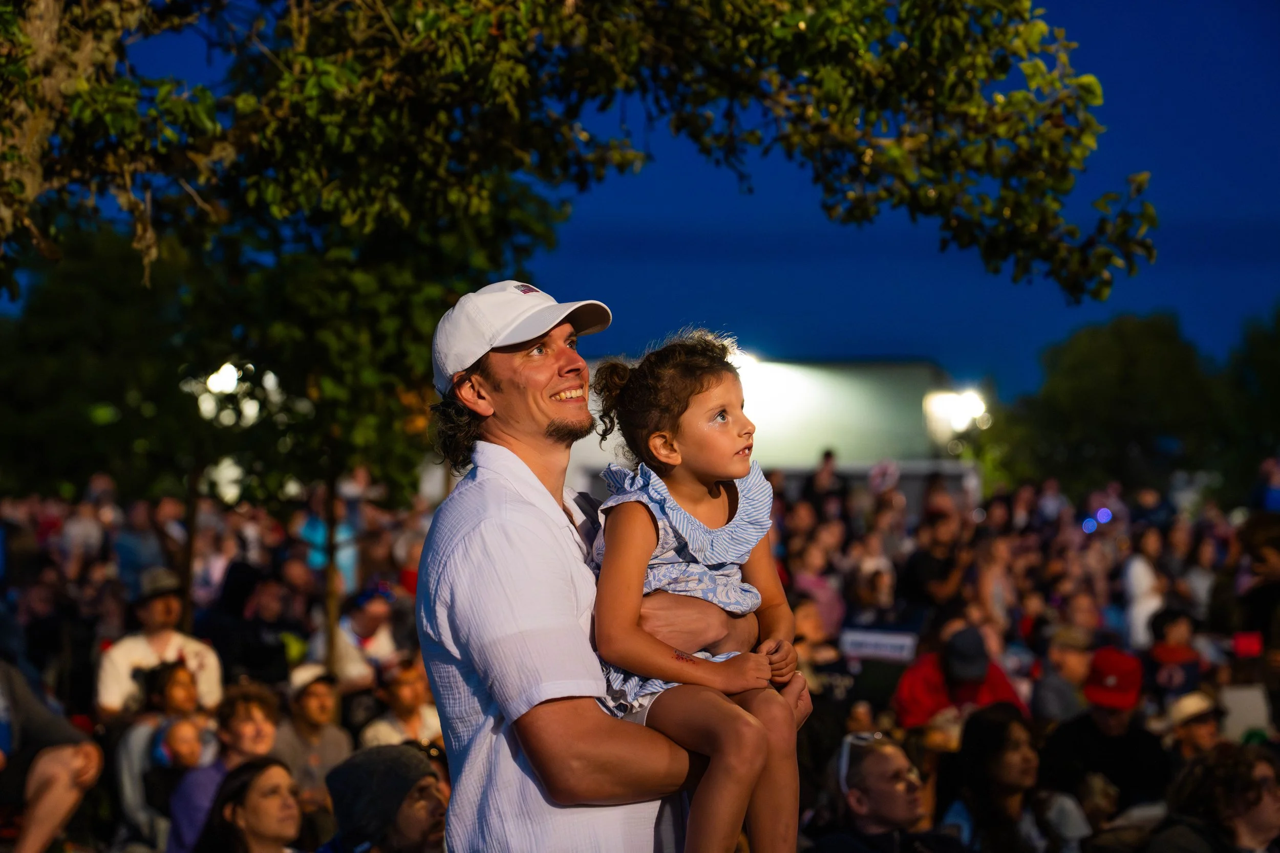 A man holding a young girl on his shoulder, watching an outdoor event at night with a crowd and trees in the background.