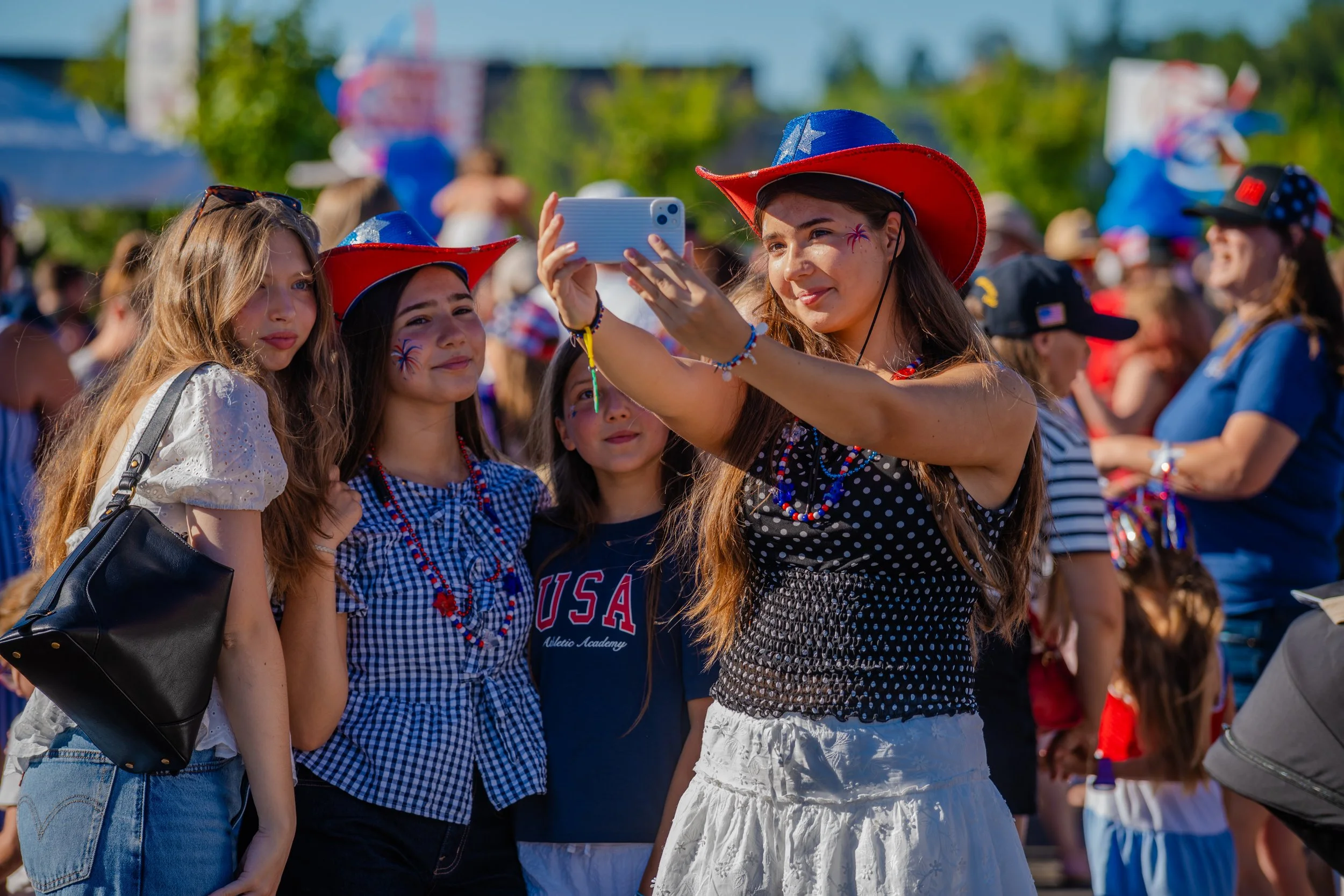A group of young women wearing red, white, and blue patriotic hats and accessories, taking a selfie at an outdoor event on a sunny day, with a crowd in the background.