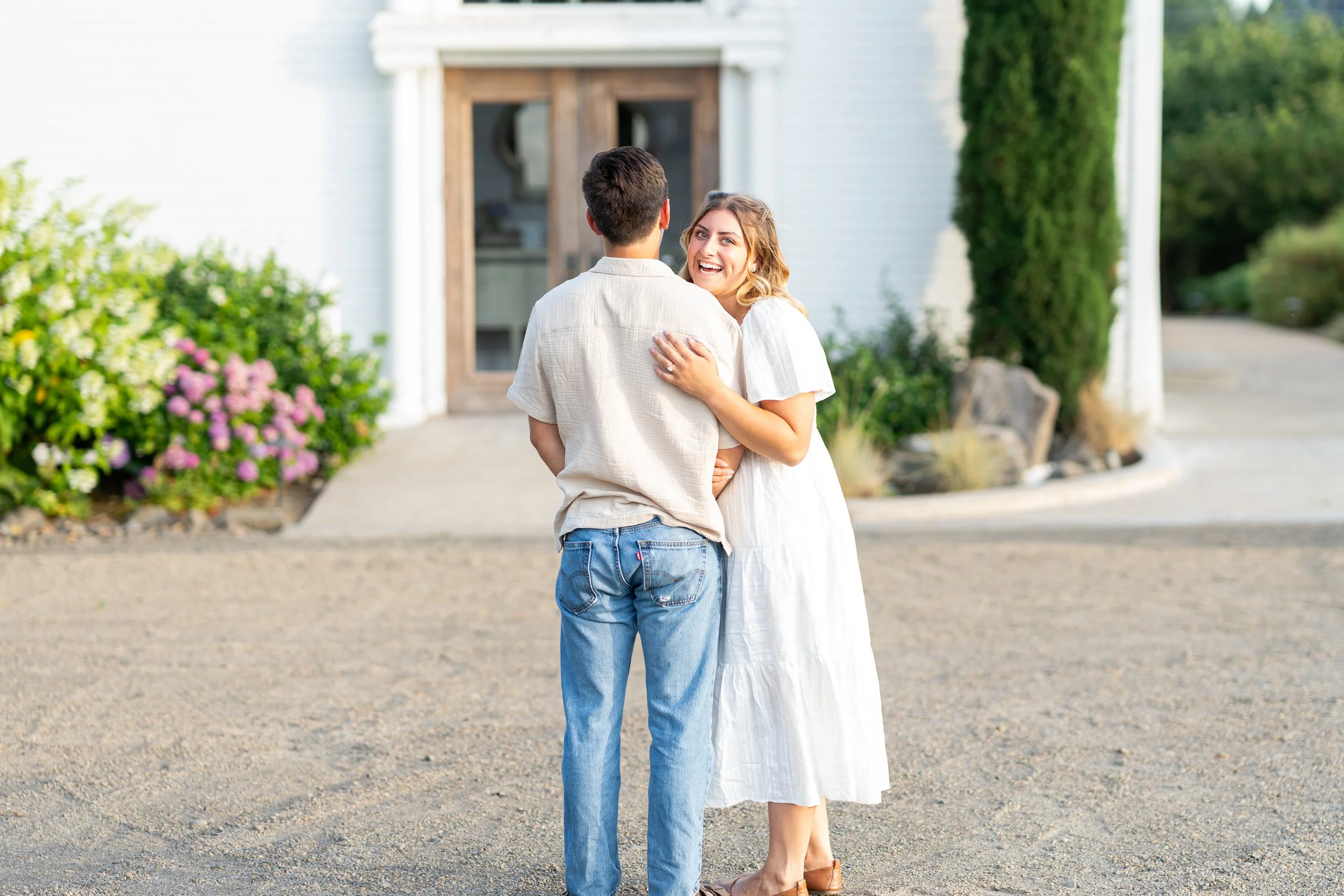 A young woman with blonde hair wearing a white dress and brown shoes hugging a young man with dark hair wearing a light-colored shirt and blue jeans outside in front of a white house with green trees and bushes.