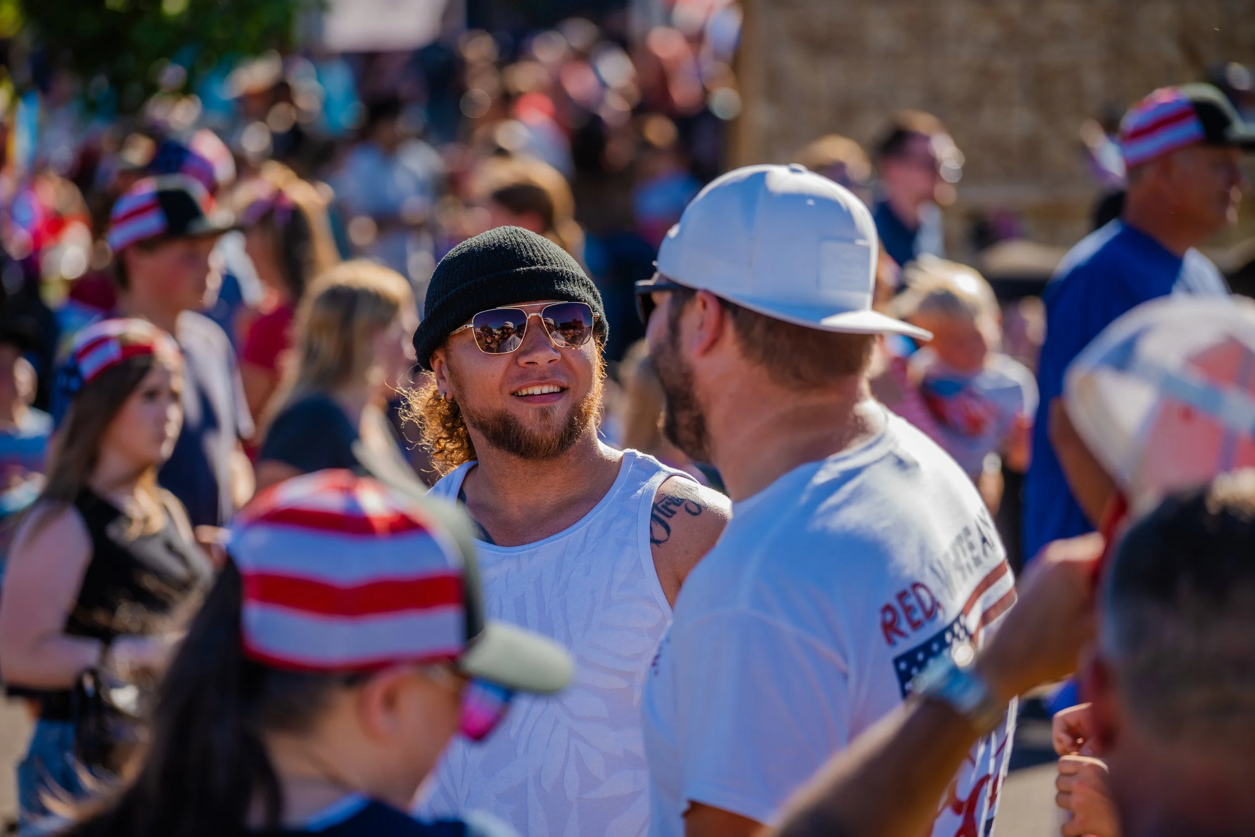 A crowd of people outdoors, some wearing patriotic clothing and hats, with a man with glasses and a black beanie in the foreground.