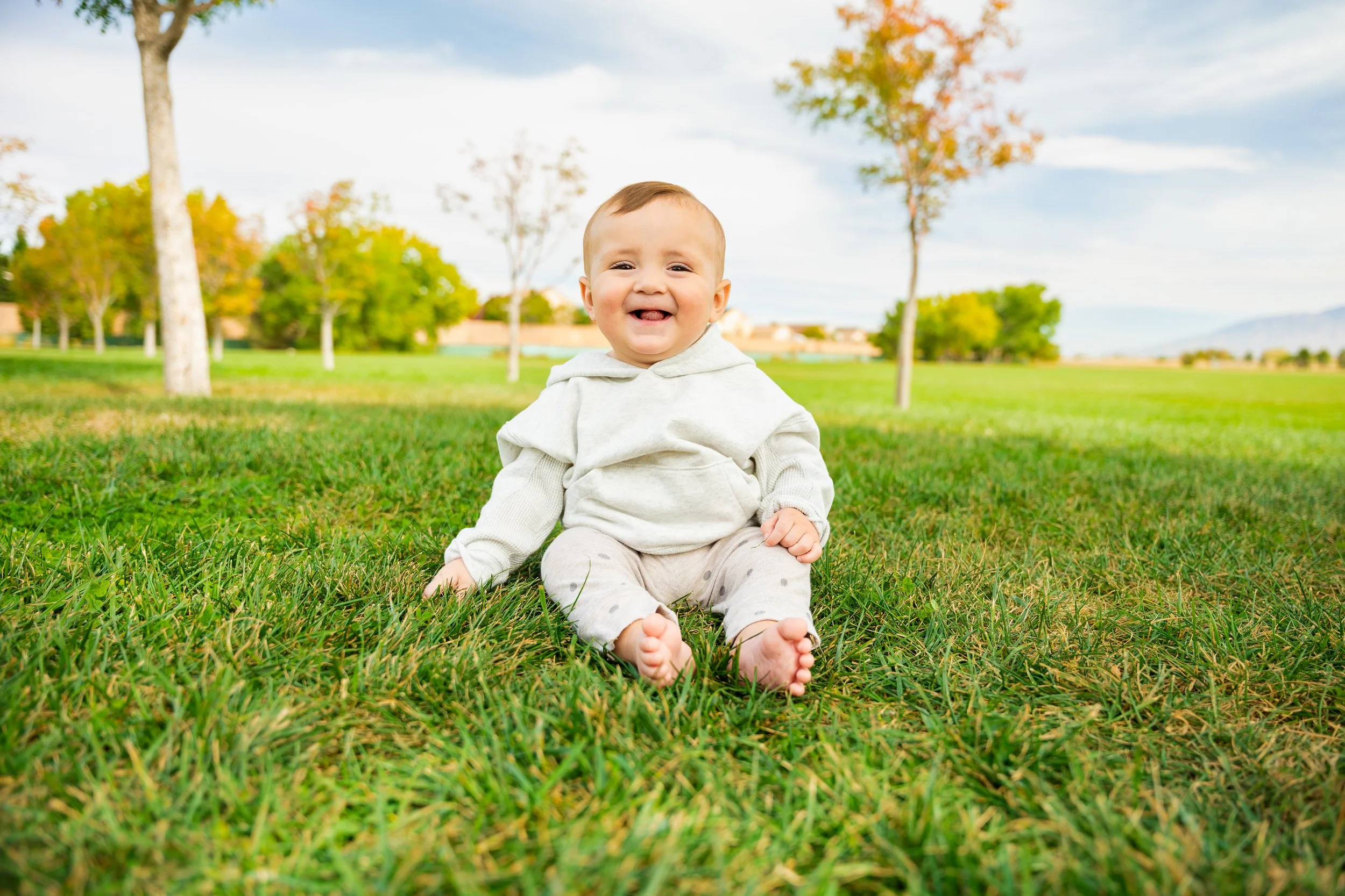 A smiling baby sitting on grass in a park with trees and blue sky in the background.