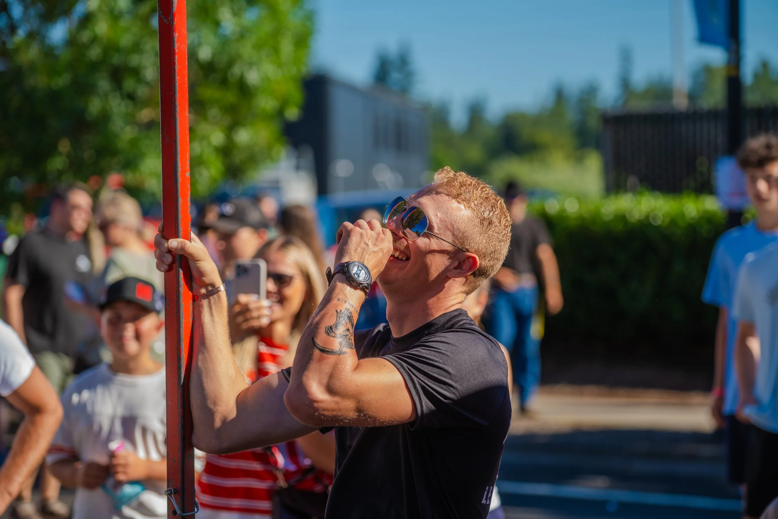 A man with tattoos and sunglasses smiling as he lifts a red pole at an outdoor event, with onlookers in the background.