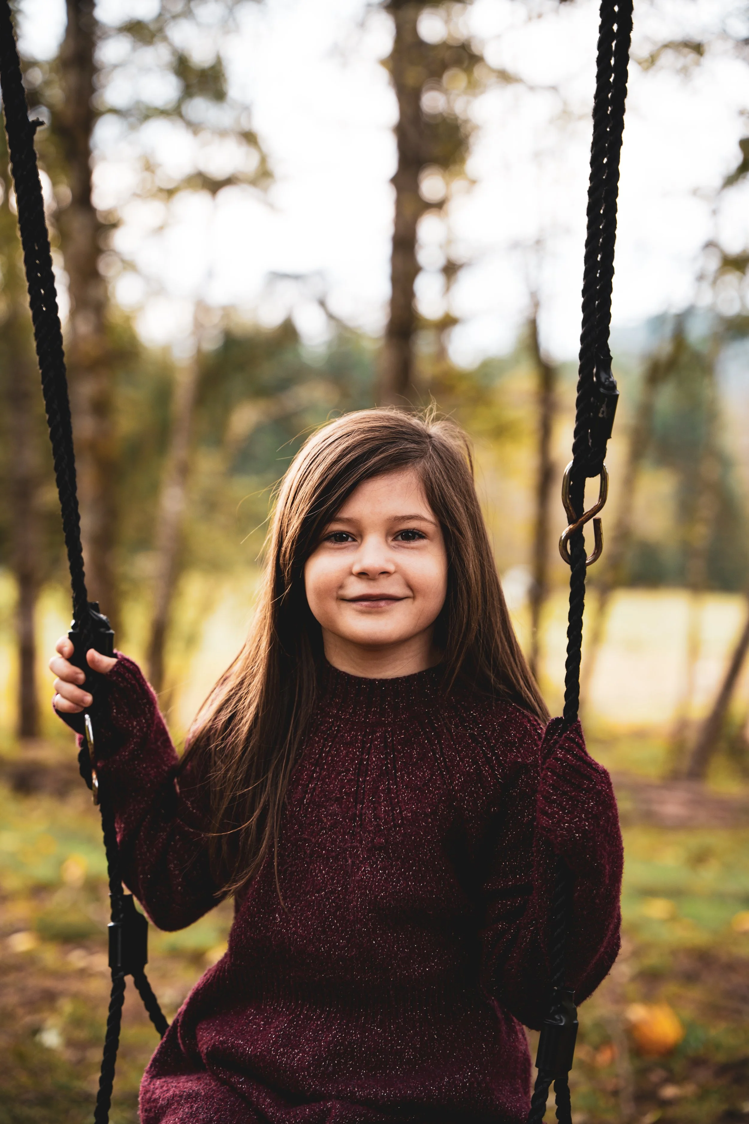 A young girl with long brown hair sits on a swing outdoors during fall, wearing a burgundy sweater, holding the ropes, and smiling at the camera with trees and fallen leaves in the background.