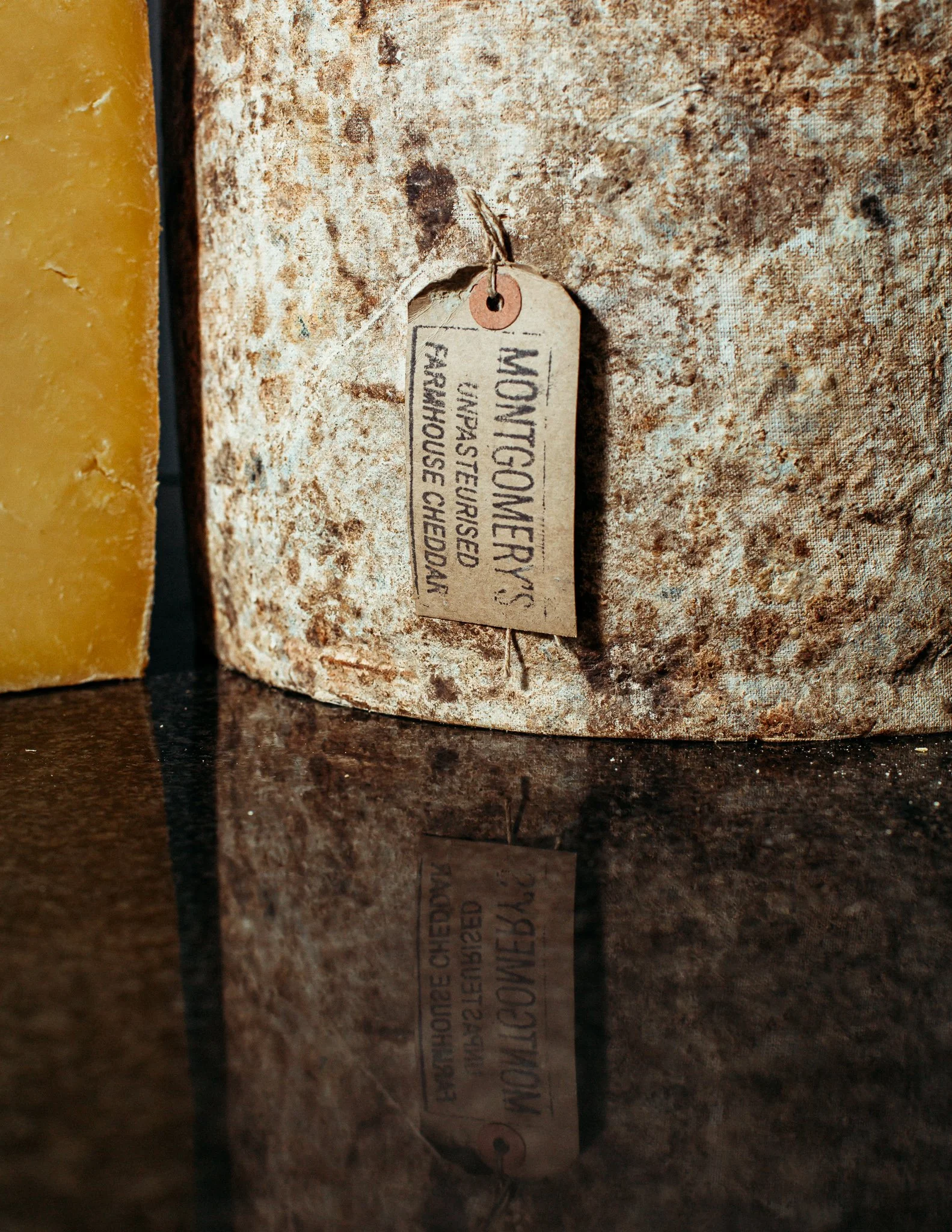 A close-up of a MONTGOMERY'S CHEDDAR. The cheese is clothbound and the label is attached with string, hanging next to the cheese on a dark, reflective surface.