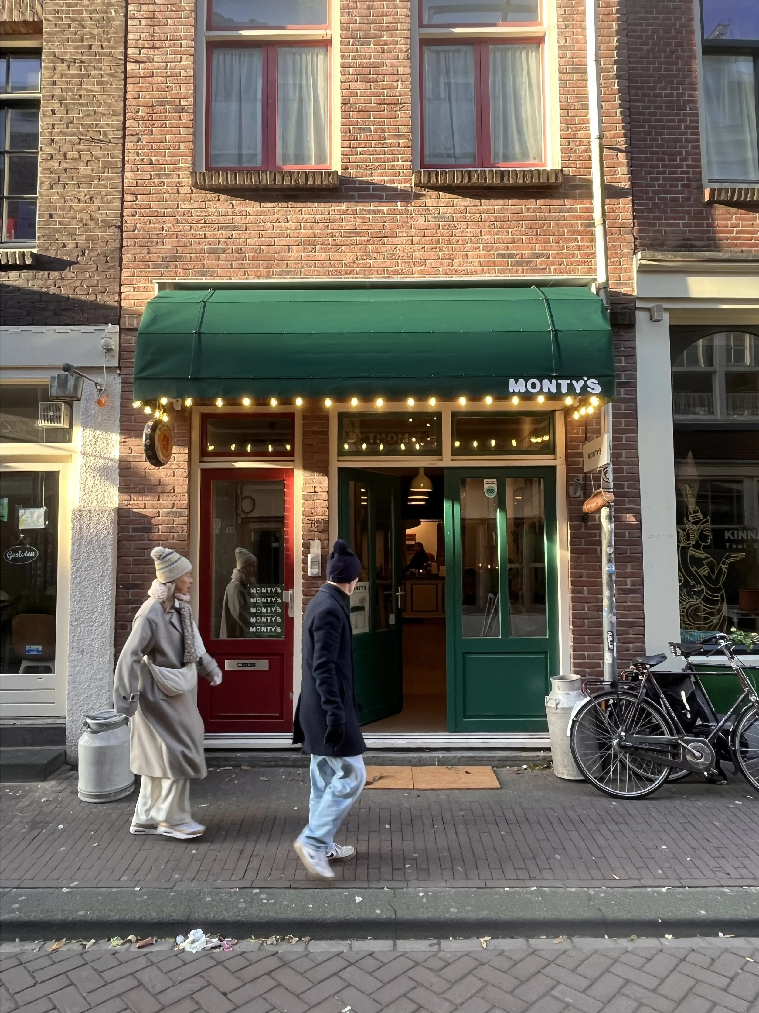 Front view of the restaurant MONTY'S with a green awning and red doors, featuring string lights; two people walking on the pavement in winter clothing.