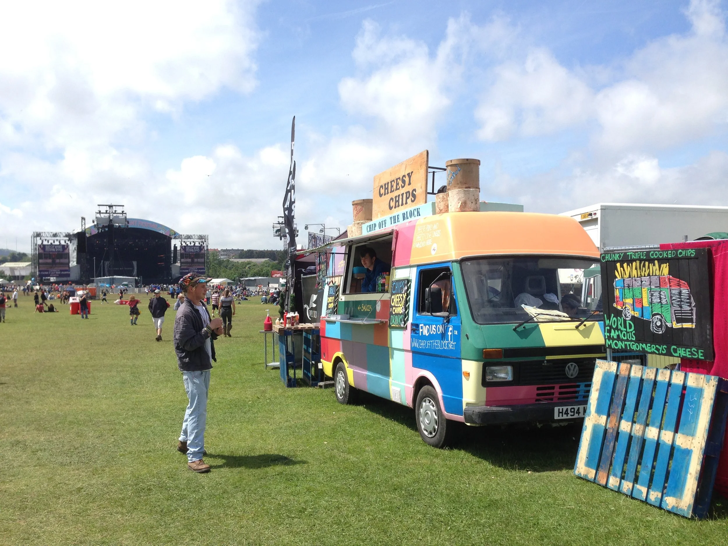 Colorful food truck selling cheesy chips, with a sign advertising 'Cheesy Chips,' at an outdoor festival. A man stands nearby holding a drink, and the background shows a large stage with people in the distance.
