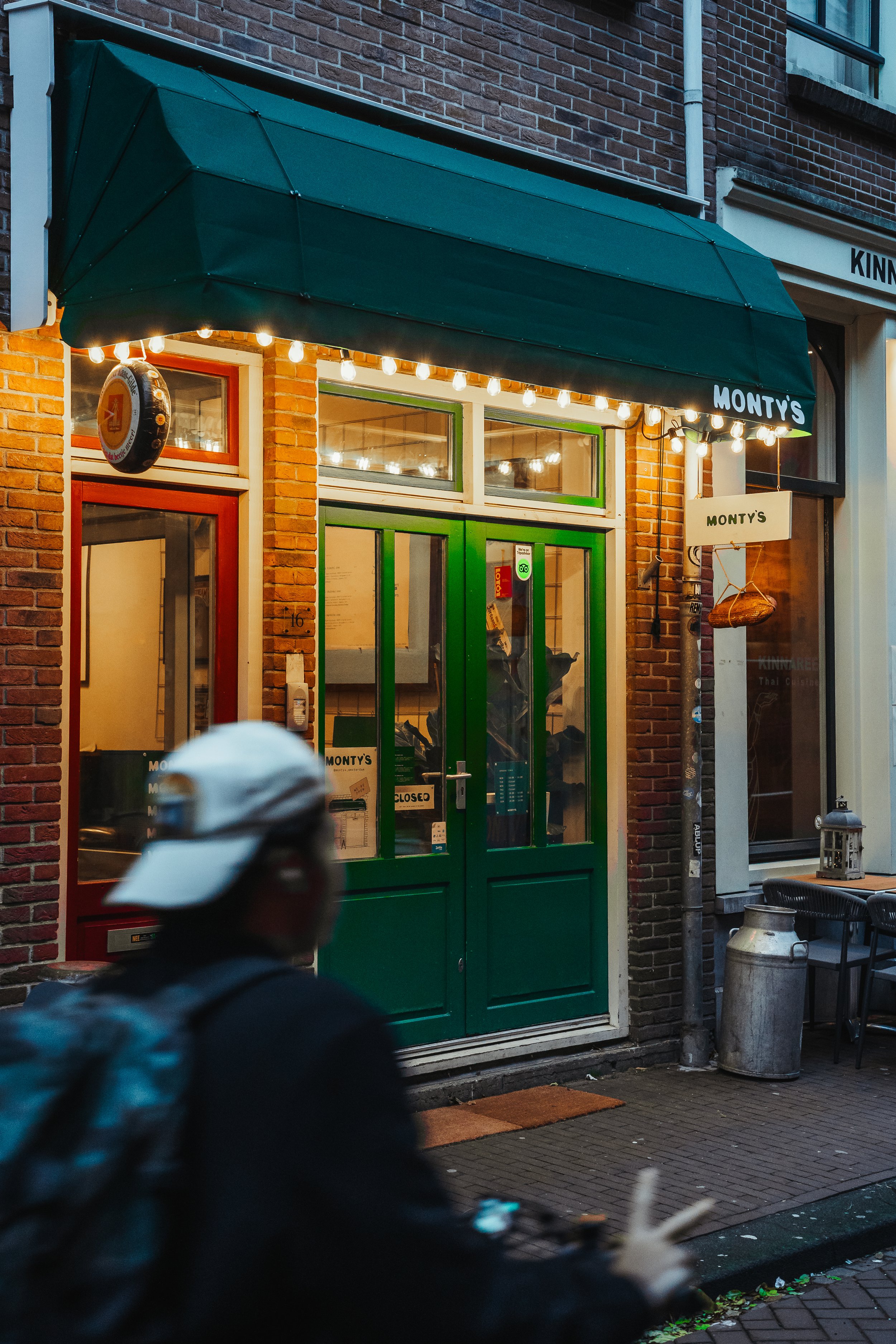 Street view of Monty's restaurant with a green door, brick exterior, and string lights, blurred person in a cap in foreground.