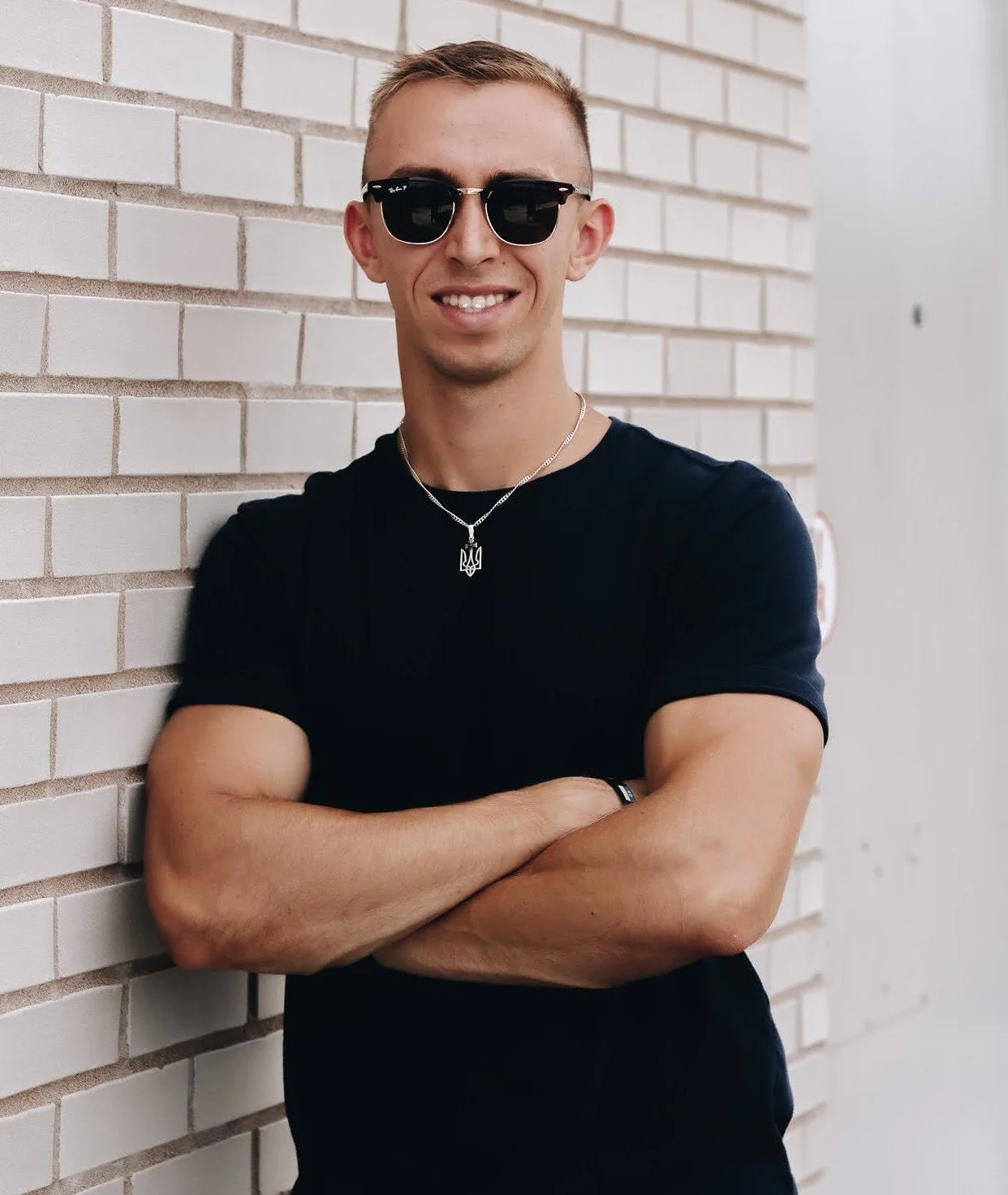 Young man with sunglasses, smiling, with arms crossed, wearing a black t-shirt and a necklace, standing against a white brick wall.