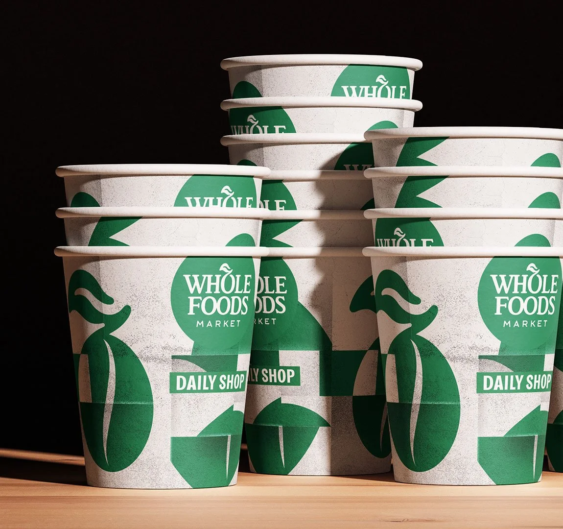 Pappbecher mit Grünem Logo von Whole Foods Market auf Holzfläche gegen schwarzen Hintergrund.