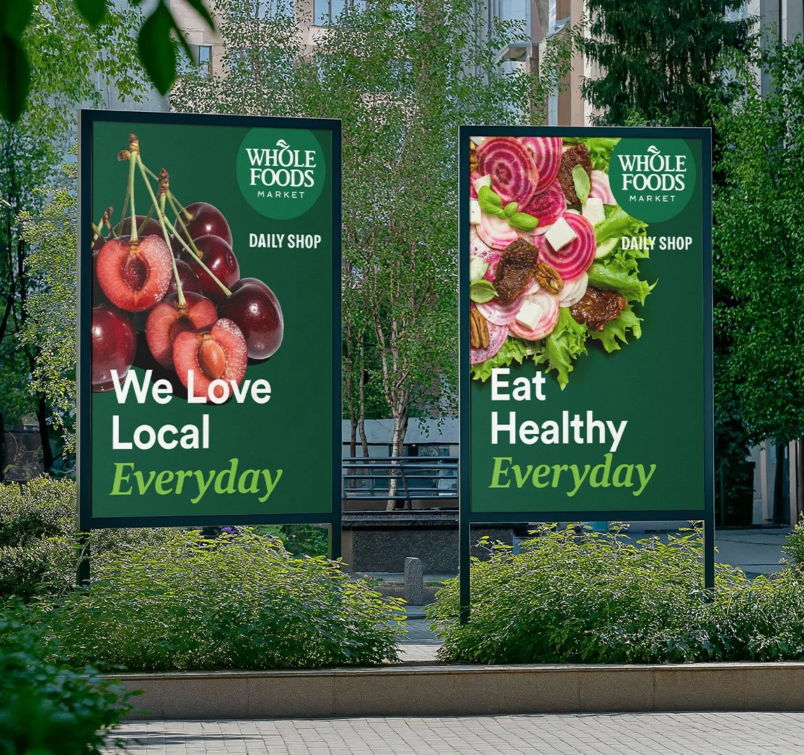 Zwei Werbetafeln im Freien für einen Naturkostladen namens Die linke Tafel zeigt Kirschen und die rechte Salat mit Radieschen, Ziegenkäse und Nüssen. Beide Tafeln tragen die Botschaft: 'We Love Local Everyday' und 'Eat Healthy Everyday'.