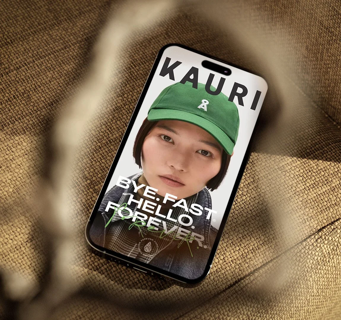 Smartphone mit einem Bild einer jungen Frau, die eine grüne Kappe trägt, auf dem Bildschirm, Text: "KAURI. BYE. FAST HELLO. FOREVER."