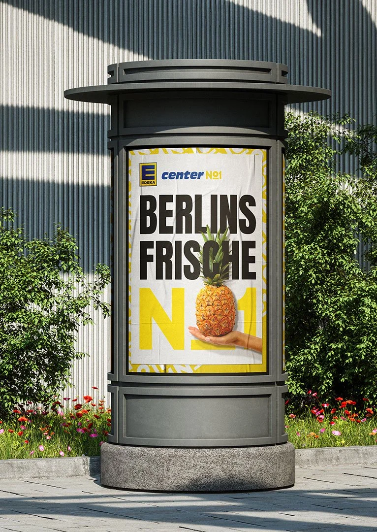 EDEKA No1 - Werbung für frische Ananas vor einem Gebäude, mit bunten Blumen und grünen Pflanzen im Hintergrund.
