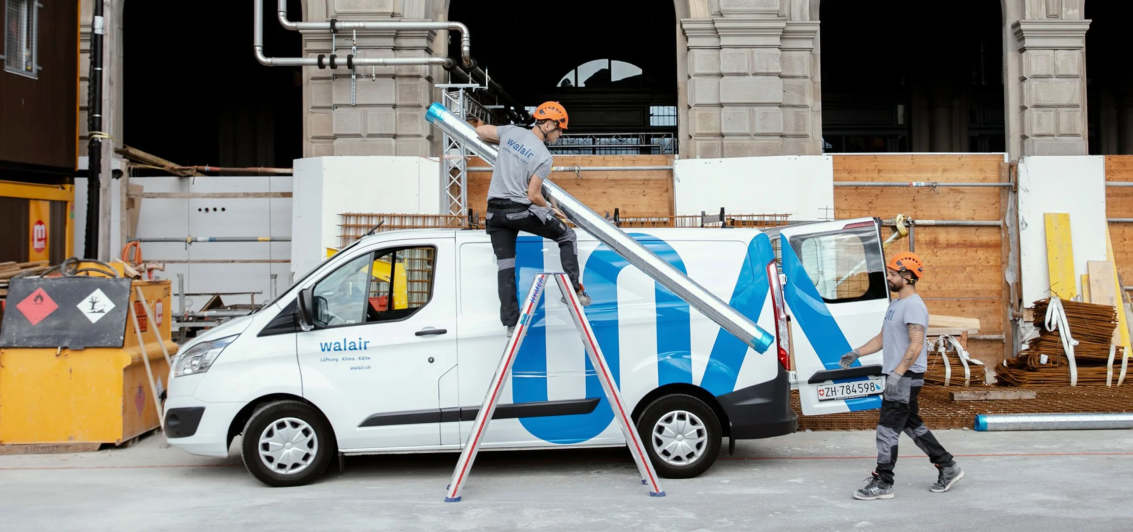 WALAIR Brand Design - Zwei Arbeiter mit Helmen arbeiten an einem weißen Firmenfahrzeug auf einer Baustelle, einer steht auf einer Leiter und hält ein Metallrohr, der andere steht neben dem Auto.