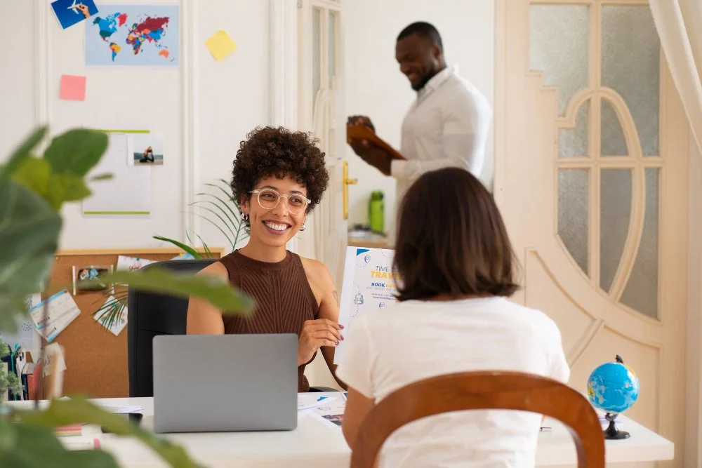 Smiling woman with curly hair and glasses sitting at a desk, showing a colorful flyer to another person in an office with a world map and colorful sticky notes on the wall.