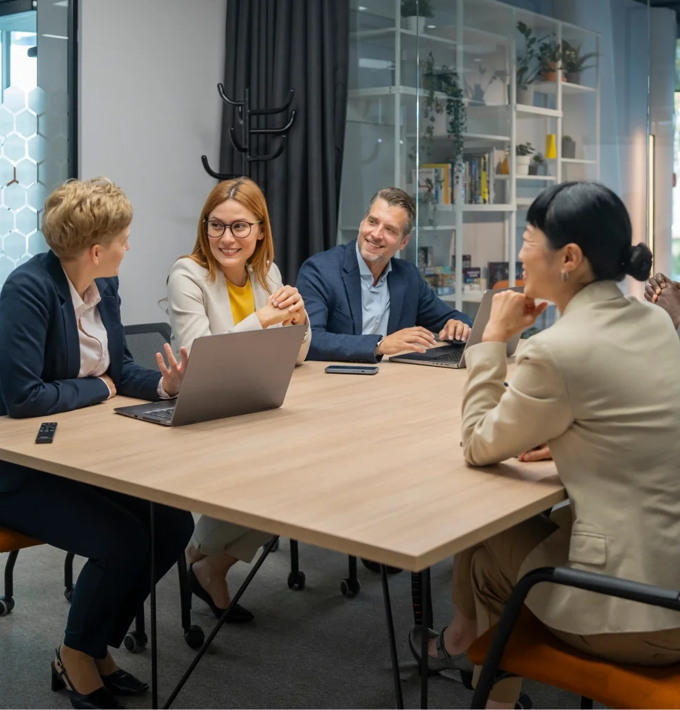 A group of four professionals in a meeting room engaged in conversation, seated around a wooden table with laptops and a remote control. They are smiling and appear to be in a discussion.