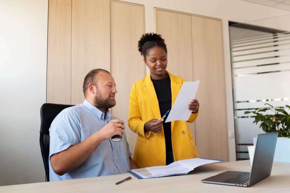 HRERDR. A man sitting at a desk next to a woman in a yellow blazer holding a piece of paper, both smiling in an office setting.