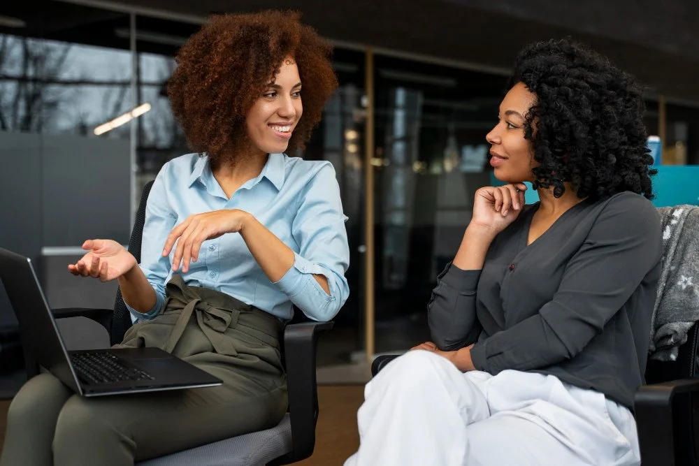 Two women sitting and talking in an office, one with curly brown hair wearing a light blue shirt, the other with curly black hair wearing a dark blouse, engaged in conversation.