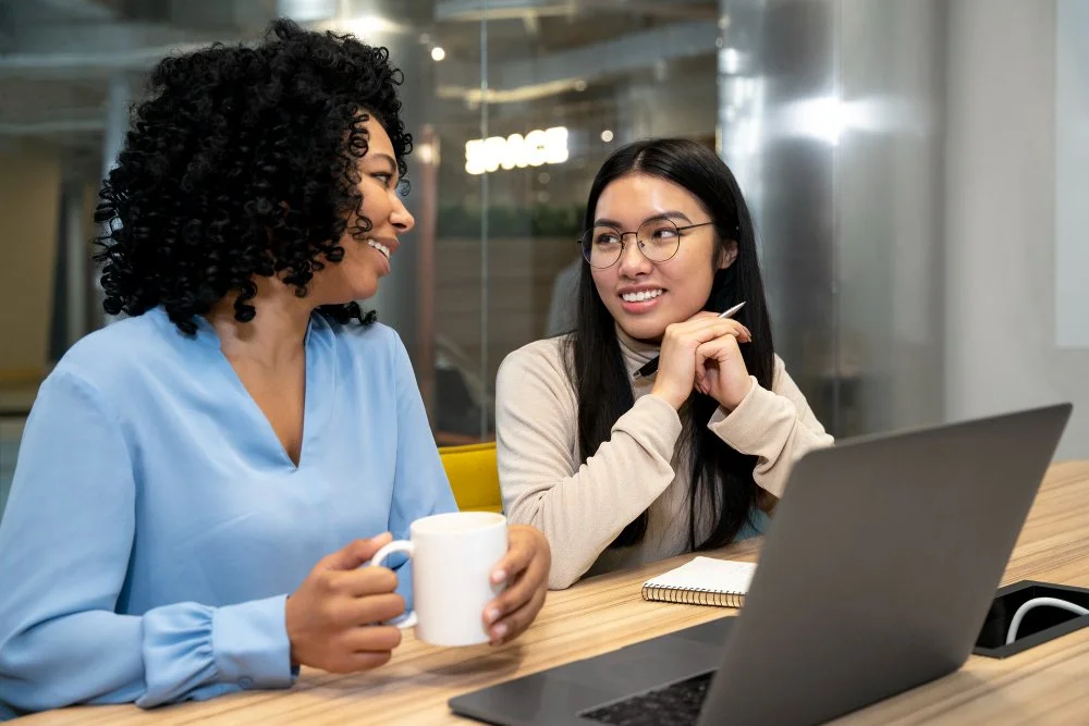 HRERDR. Two women having a discussion at a wooden table with a laptop, notebook, and mug in an office setting.