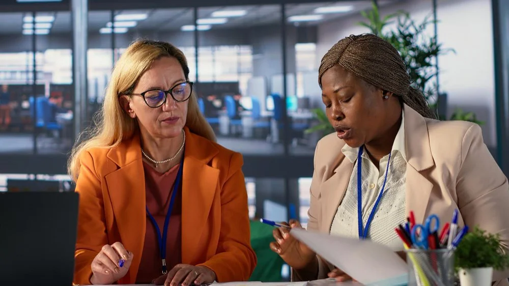 Two women in business attire having a serious discussion in a modern office with glass walls and plants.