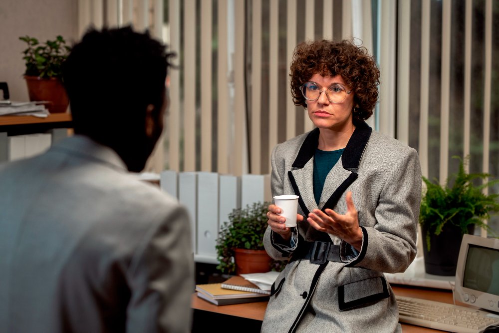 A woman with curly hair, glasses, and a gray blazer is having a conversation with a man in an office setting, holding a white cup.