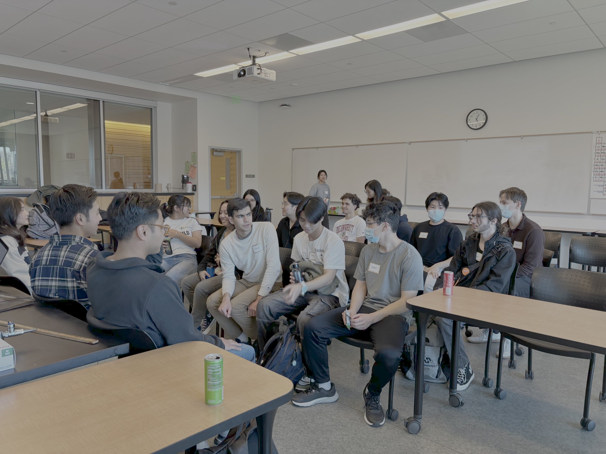 A diverse group of students sitting in a classroom, some wearing masks, engaged in a discussion or listening to a speaker at the front of the room.