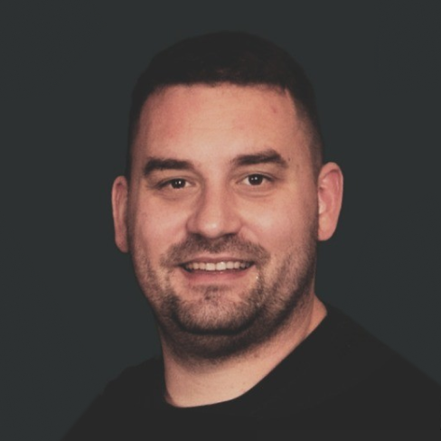 A man with short dark hair, light skin, and a beard smiling in front of a dark background.