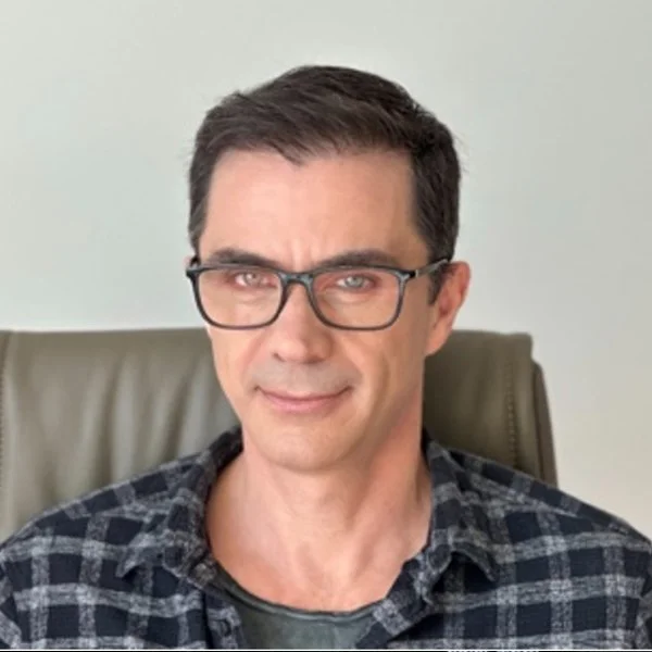 Man with dark hair and glasses sitting on a leather chair, wearing a plaid shirt and smiling slightly.