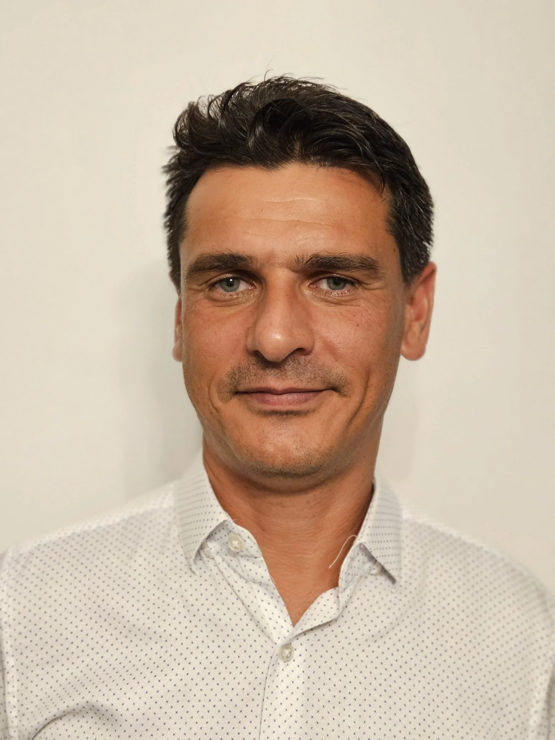 A man with dark hair and blue eyes, wearing a white button-up shirt with small polka dots, standing against a plain white background.