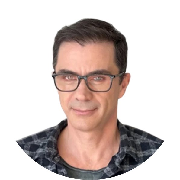 Man with dark hair and glasses sitting on a leather chair, wearing a plaid shirt and smiling slightly.